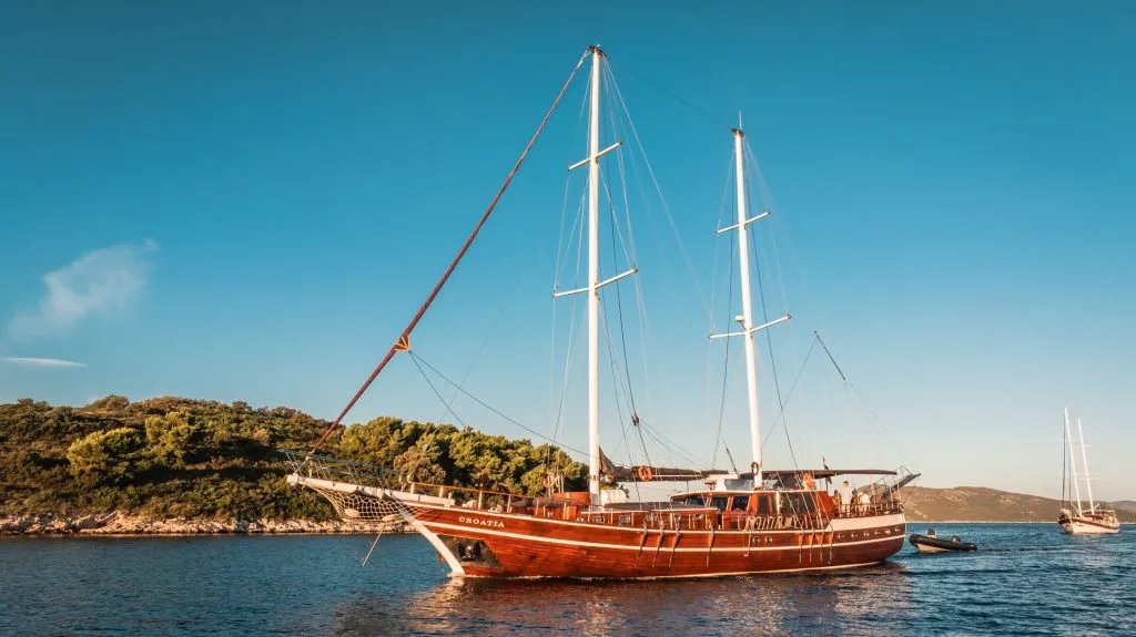 A large wooden sailboat with two masts is anchored in calm blue water near a tree-covered island under a clear sky. Another sailboat is visible in the background.