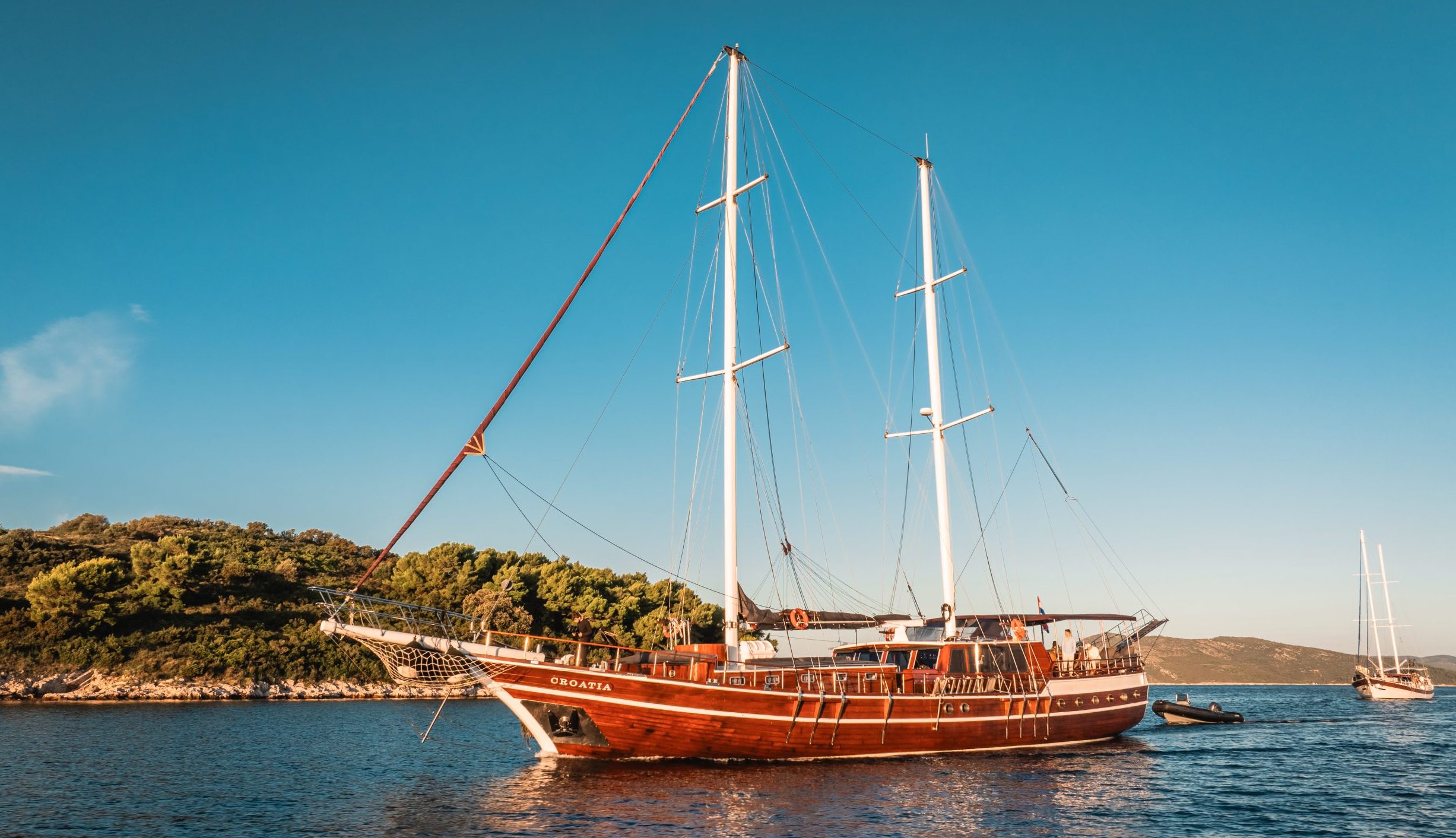 A large wooden sailboat with two masts is anchored in calm blue water near a tree-covered island under a clear sky. Another sailboat is visible in the background.