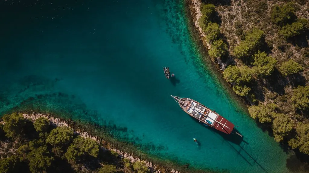 Aerial view of a large sailboat anchored in clear turquoise water near a forested coastline, with two smaller boats nearby and green trees lining the rocky shore.