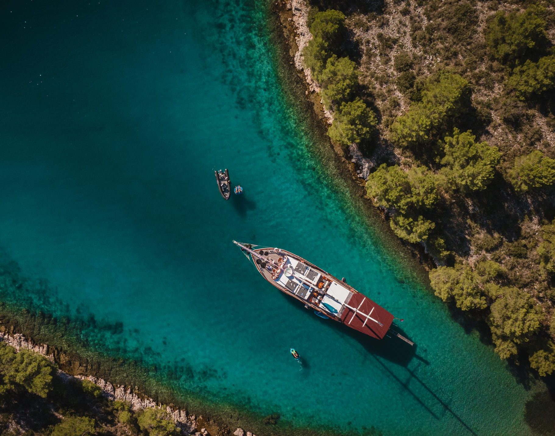 Aerial view of a large sailboat anchored in clear turquoise water near a forested coastline, with two smaller boats nearby and green trees lining the rocky shore.