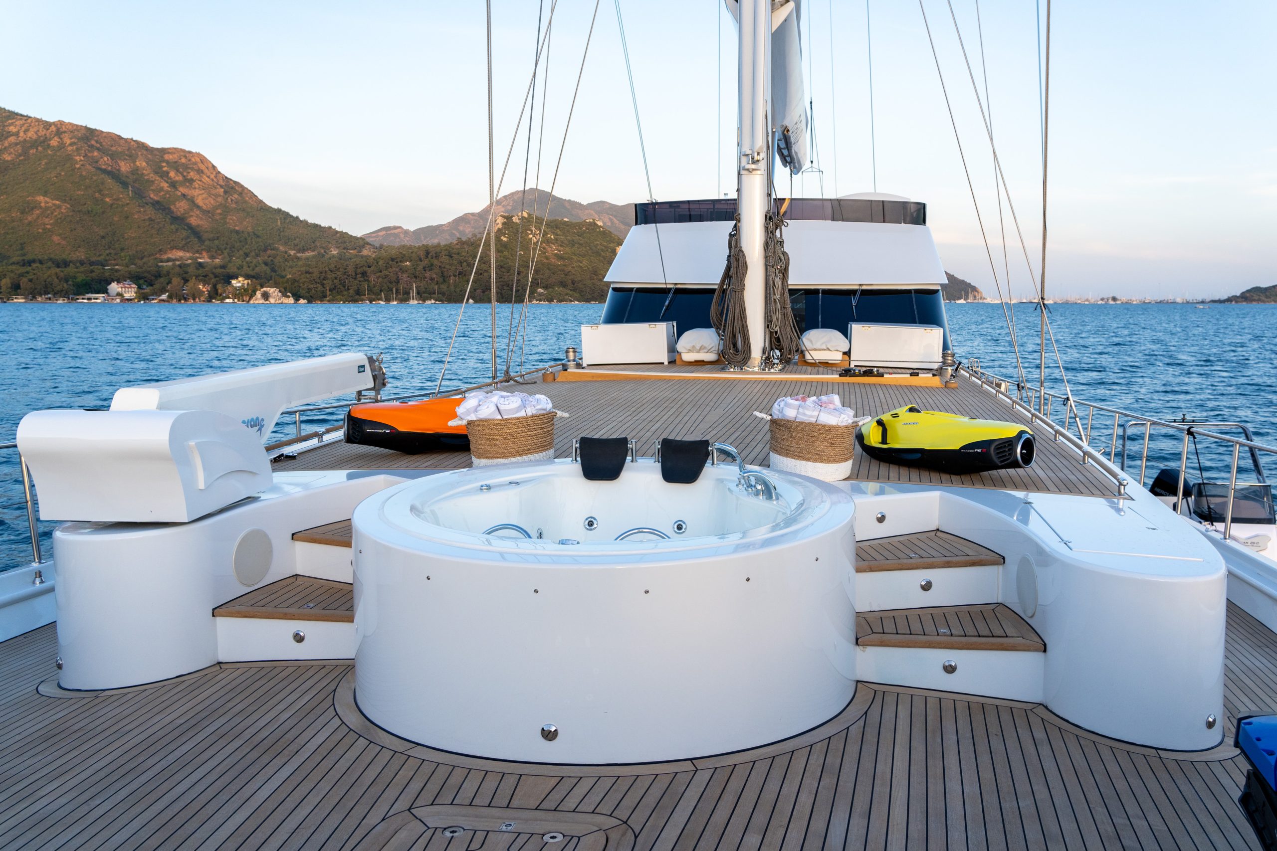 GOOD LIFE A luxurious yacht deck features a circular jacuzzi with three headrests, surrounded by teak steps. The yacht is on calm water, with mountains and greenery in the background under a clear sky.