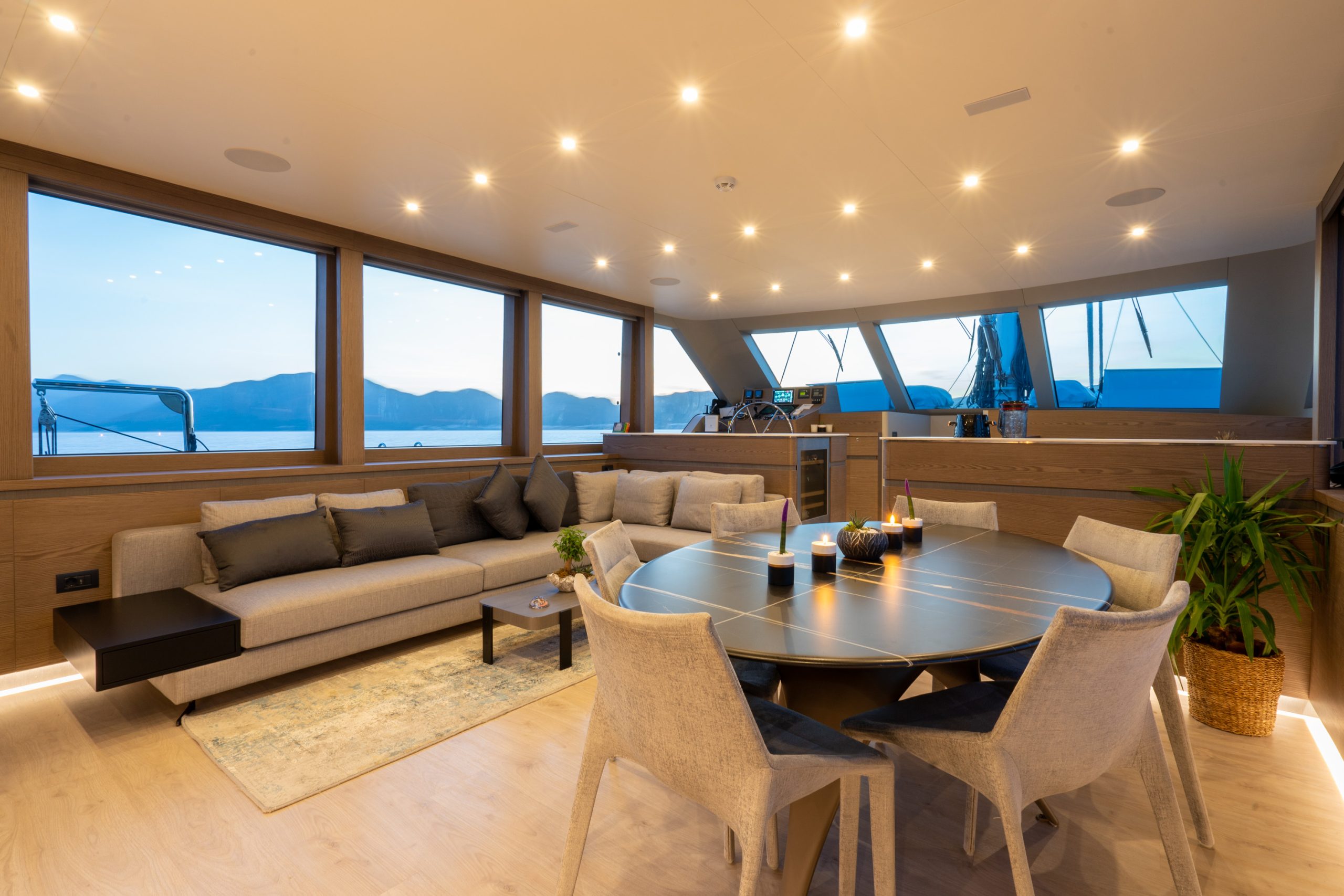 GOOD LIFE Modern yacht interior featuring a round dining table with six chairs, a gray sectional sofa, large windows with water and mountain views, wooden floors, and soft, warm lighting. A potted plant sits near the table.