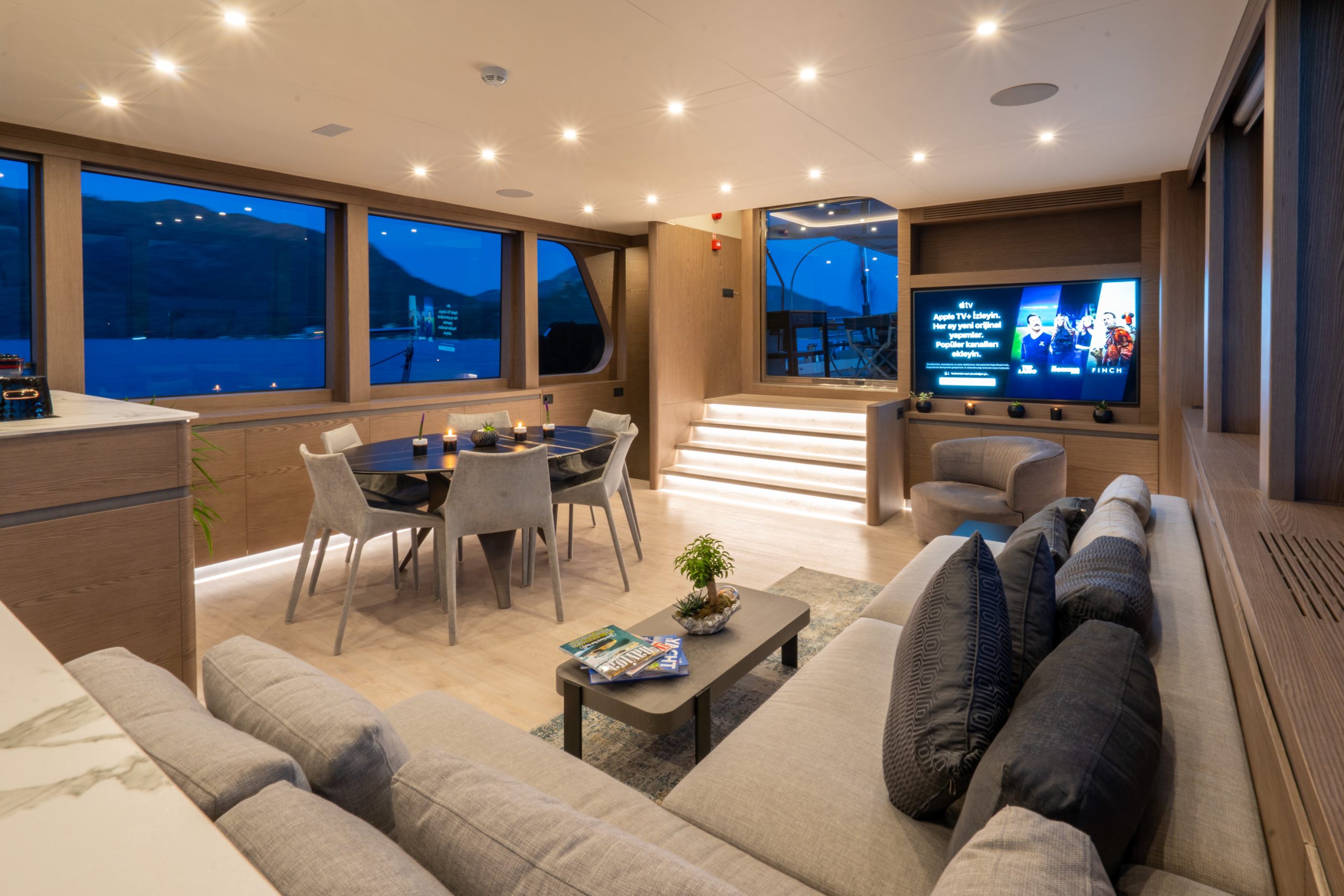 GOOD LIFE A modern yacht interior with a gray sectional sofa, dining table, and a large TV on the wall. Warm lighting, large windows, and steps leading to a glass door create an inviting, luxurious atmosphere.