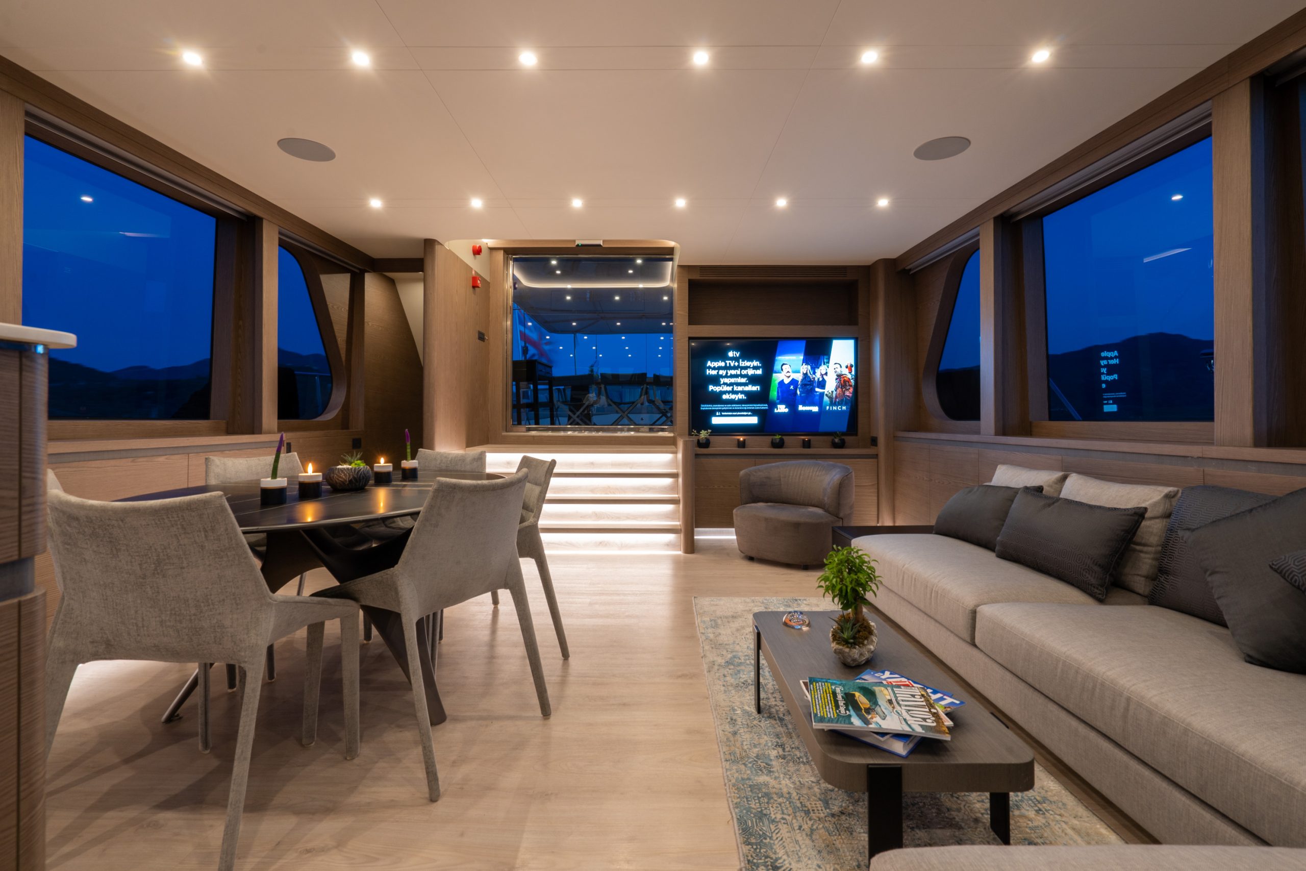 GOOD LIFE A modern yacht interior at dusk features a dining table with chairs, a long sofa, a coffee table with magazines, and a TV on the wall. Large windows showcase the dark blue evening sky outside.