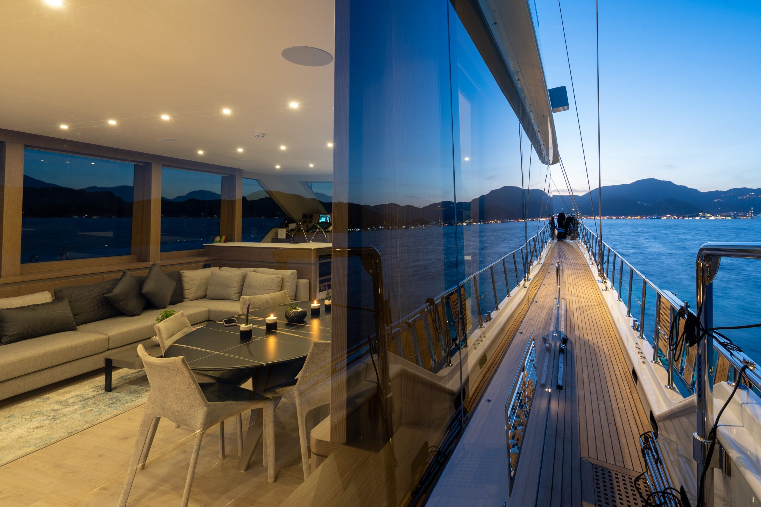 GOOD LIFE A luxury yacht deck at sunset, showing a sleek wooden walkway beside large glass windows that reflect the scenic water and distant mountains. The interior features modern seating and soft lighting.