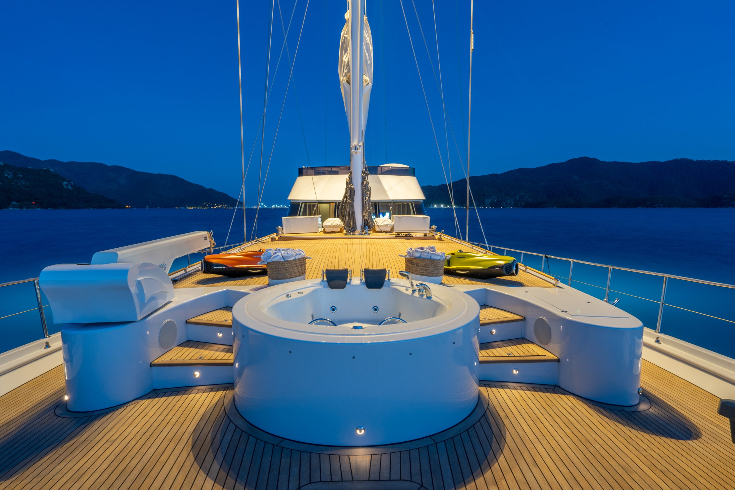 GOOD LIFE A luxurious yacht deck at dusk features a round hot tub, sun loungers, and sleek seating areas, with calm blue water and distant hills under a deep blue sky in the background.