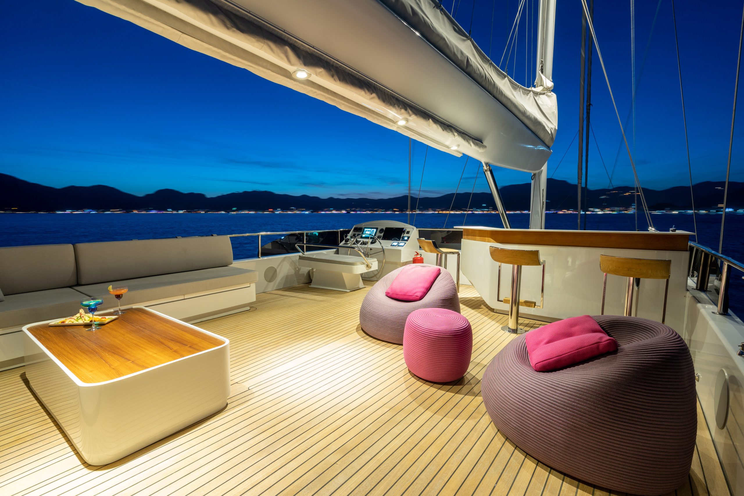 GOOD LIFE Luxury yacht deck at dusk with bean bag chairs, a small table, cushioned benches, and two bar stools. Calm sea and distant lights are visible under a deep blue sky in the background.
