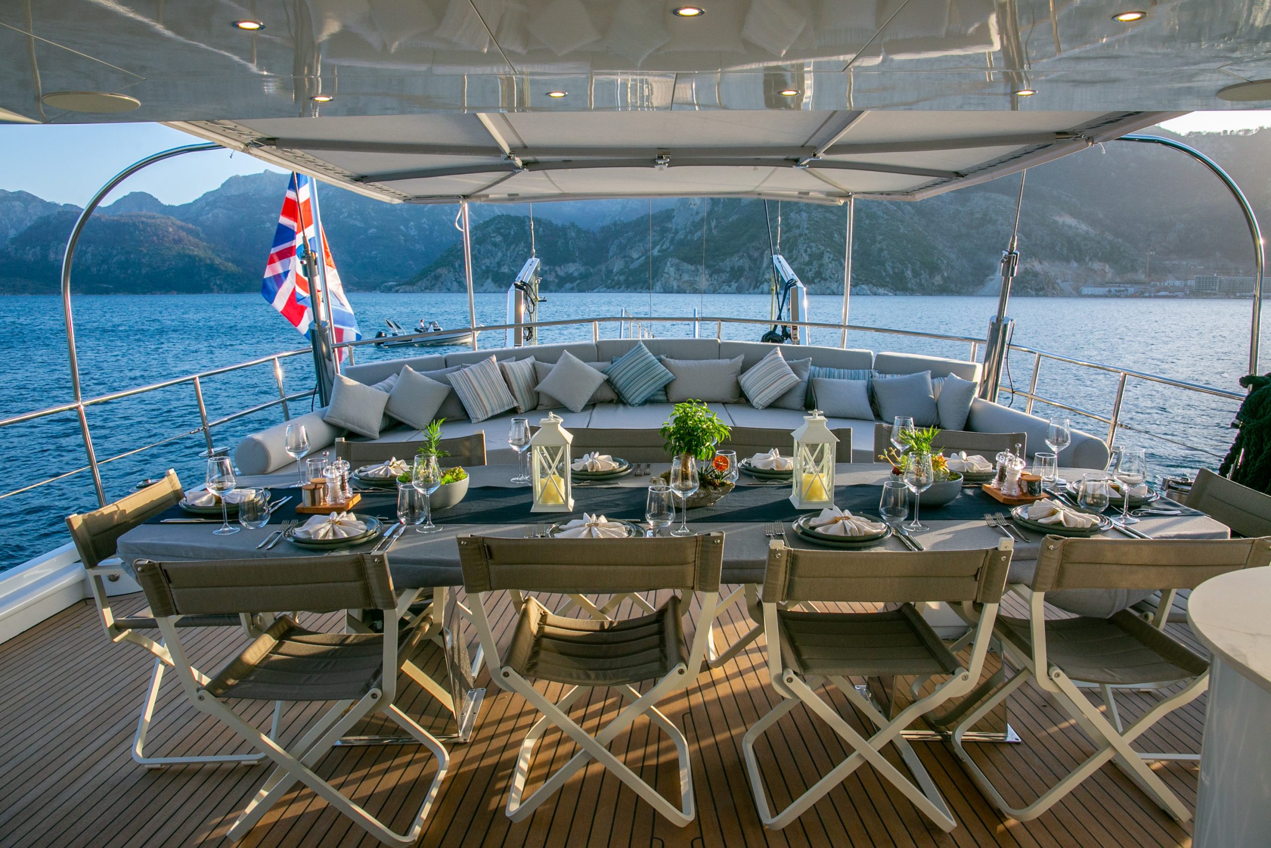 GOOD LIFE A large, elegant outdoor dining table is set on the deck of a yacht, with cushioned seating, lanterns, and place settings. The yacht is on blue water, surrounded by mountains, under a canopy.
