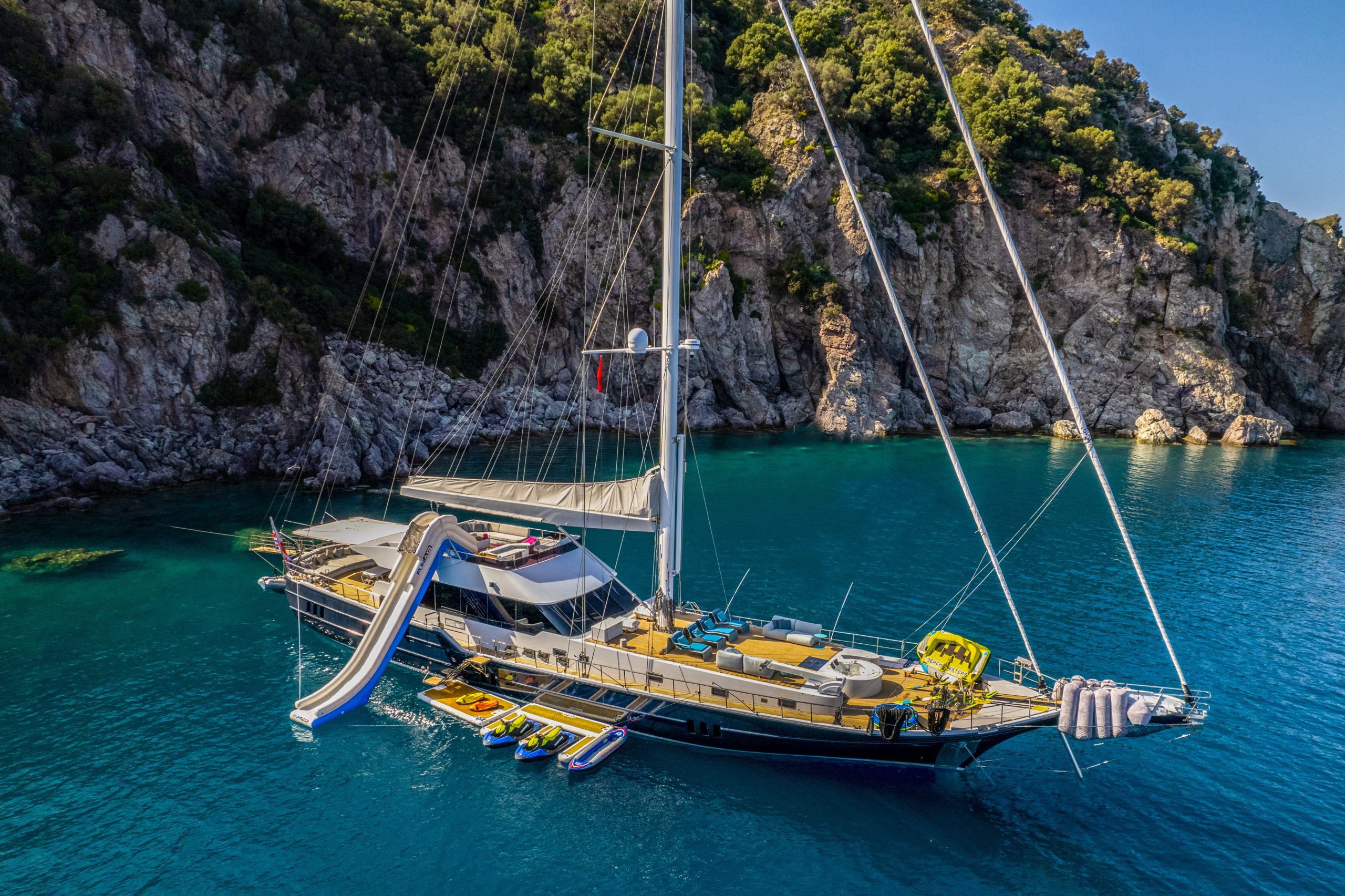 GOOD LIFE A large sailboat with a waterslide and several paddleboards floats on clear blue water near a rocky, tree-lined coastline.