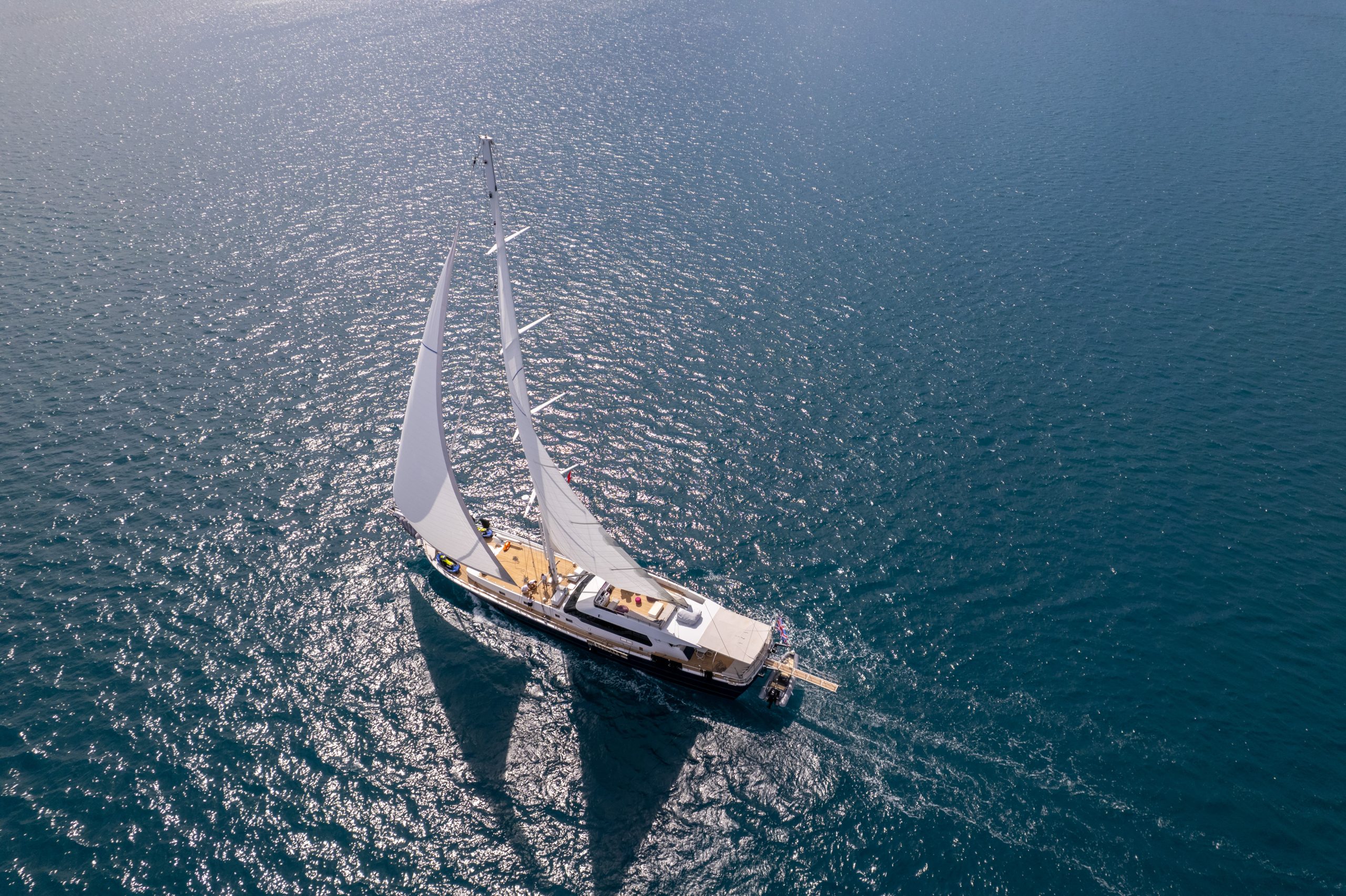 GOOD LIFE Aerial view of a sailboat with white sails gliding across calm, deep blue water under bright sunlight, leaving a gentle wake behind.