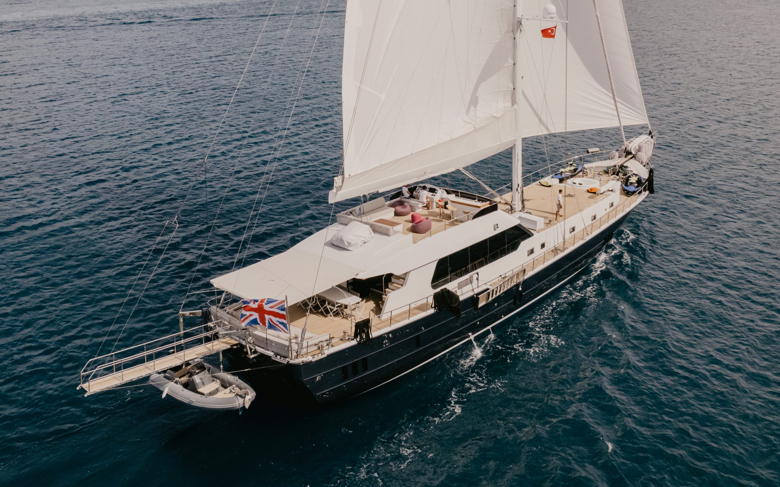 GOOD LIFE A large sailboat with white sails and a British flag is cruising on calm blue ocean water, seen from above. The yacht has a spacious deck with seating and a dinghy at the back.