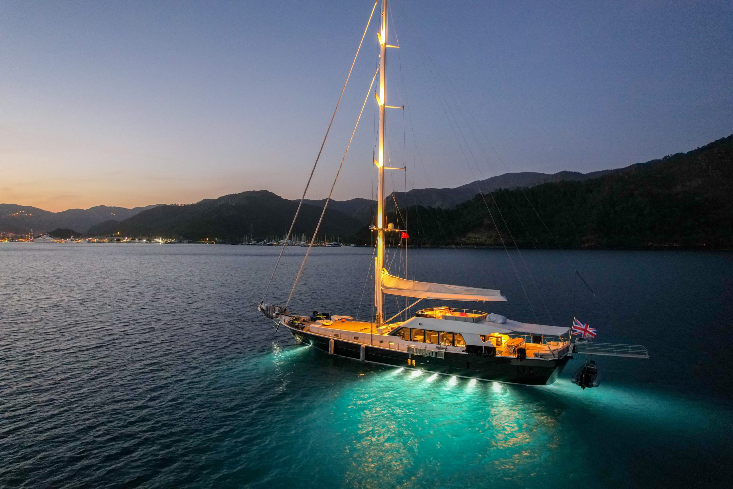 GOOD LIFE A large sailboat with illuminated deck lights floats on calm blue water at dusk, near a mountainous shoreline with city lights visible in the distance.