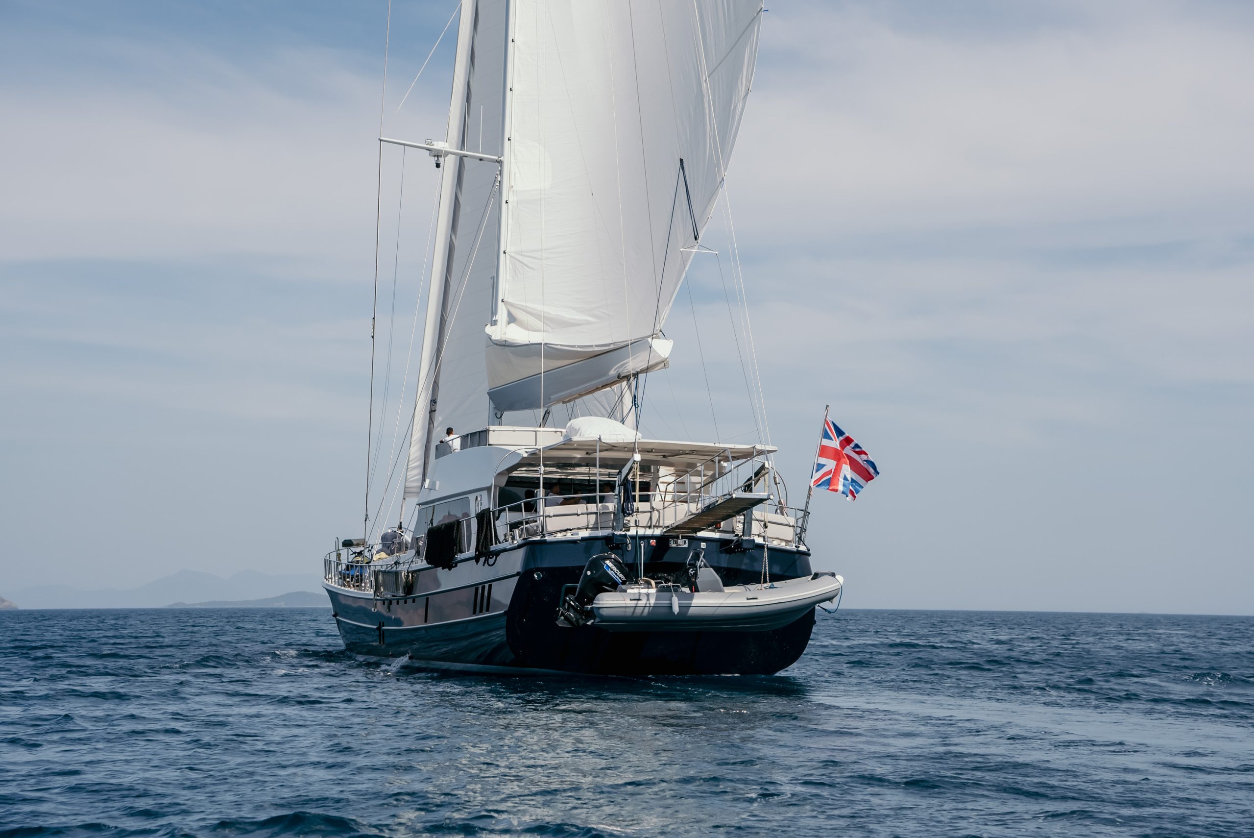 GOOD LIFE A large sailboat with white sails and a British flag at the stern travels on calm blue water under a cloudy sky. A small inflatable boat is attached to the back of the yacht.