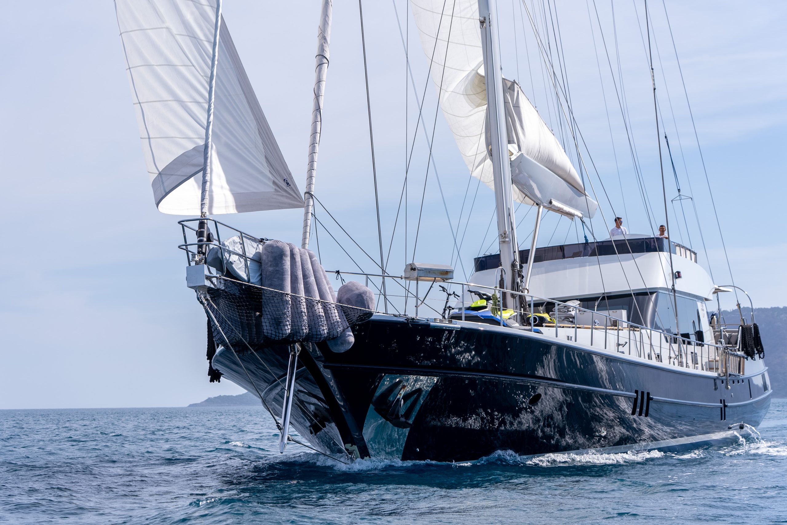 GOOD LIFE A large sailing yacht with white sails is cruising on calm blue water. Two people are visible on the upper deck, and fenders hang over the side of the sleek black hull. An island is in the background under a clear sky.
