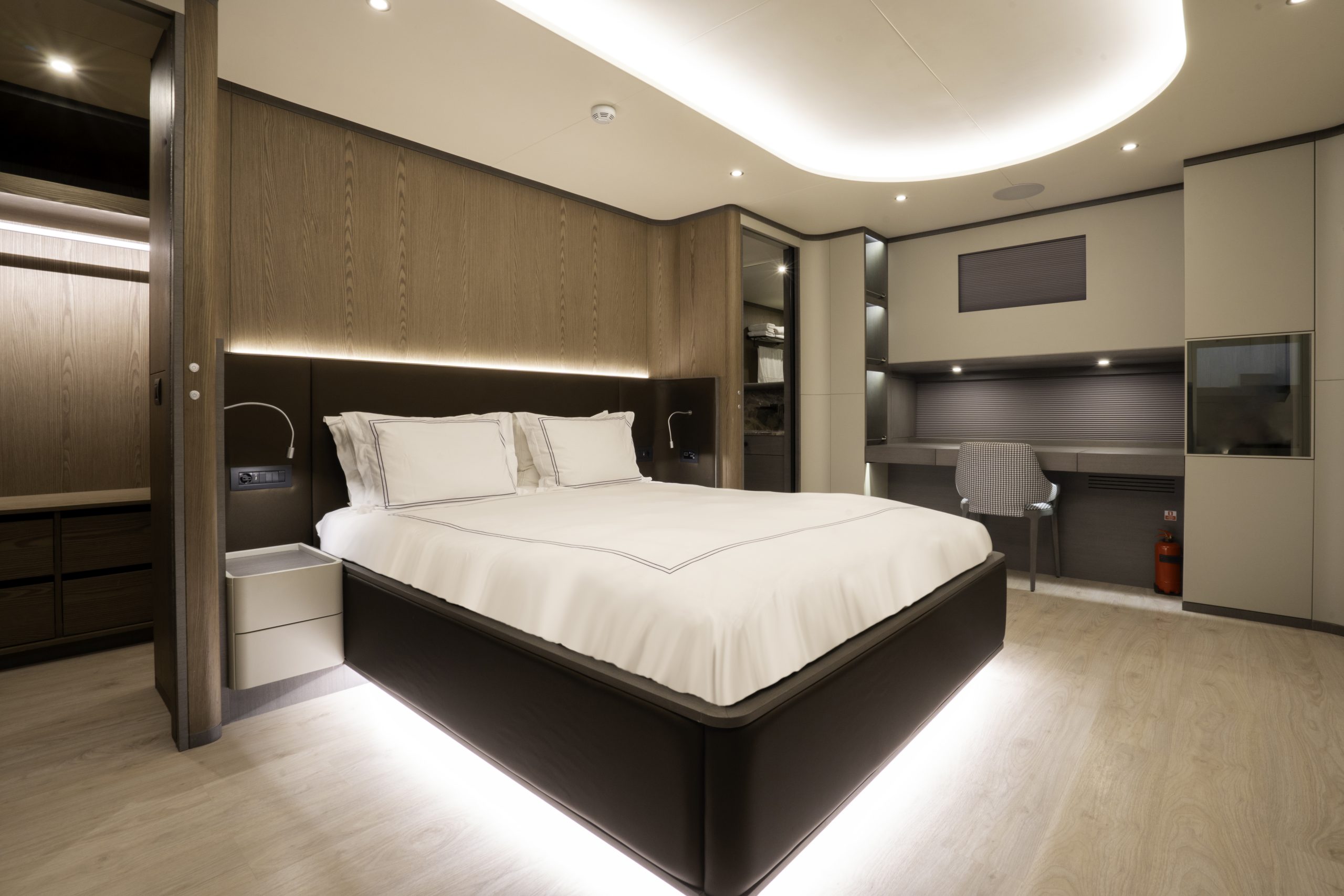 GOOD LIFE A modern, spacious bedroom with a large bed, white linens, wood paneling, built-in shelves, a work desk with a chair, and soft under-bed lighting, creating a cozy and minimalist atmosphere.