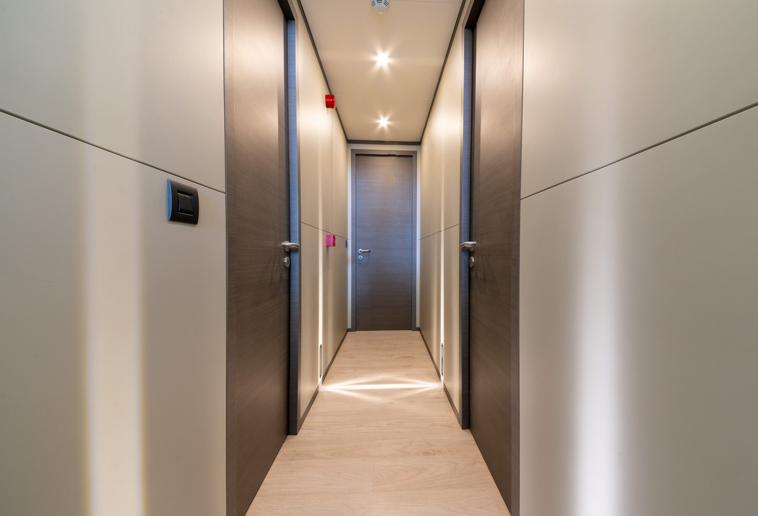 GOOD LIFE A modern, narrow hallway with light wood flooring, cream-colored walls, and dark wooden doors on both sides, leading to a closed door at the end. The space is brightly lit by ceiling lights.