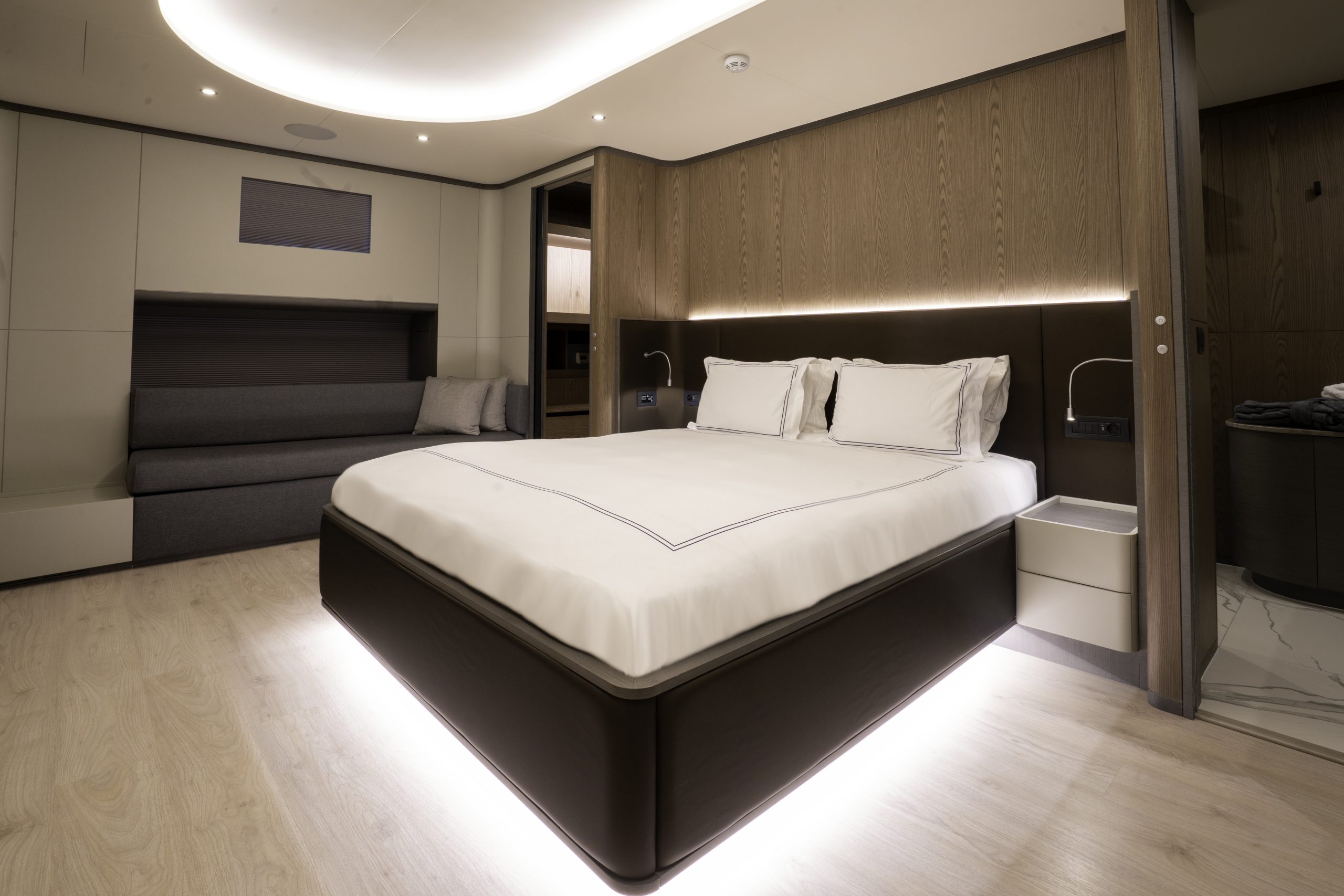 GOOD LIFE A modern, minimalistic bedroom with a large bed featuring white bedding, built-in headboard lighting, light wood floors, neutral walls, and a gray sofa against the wall.