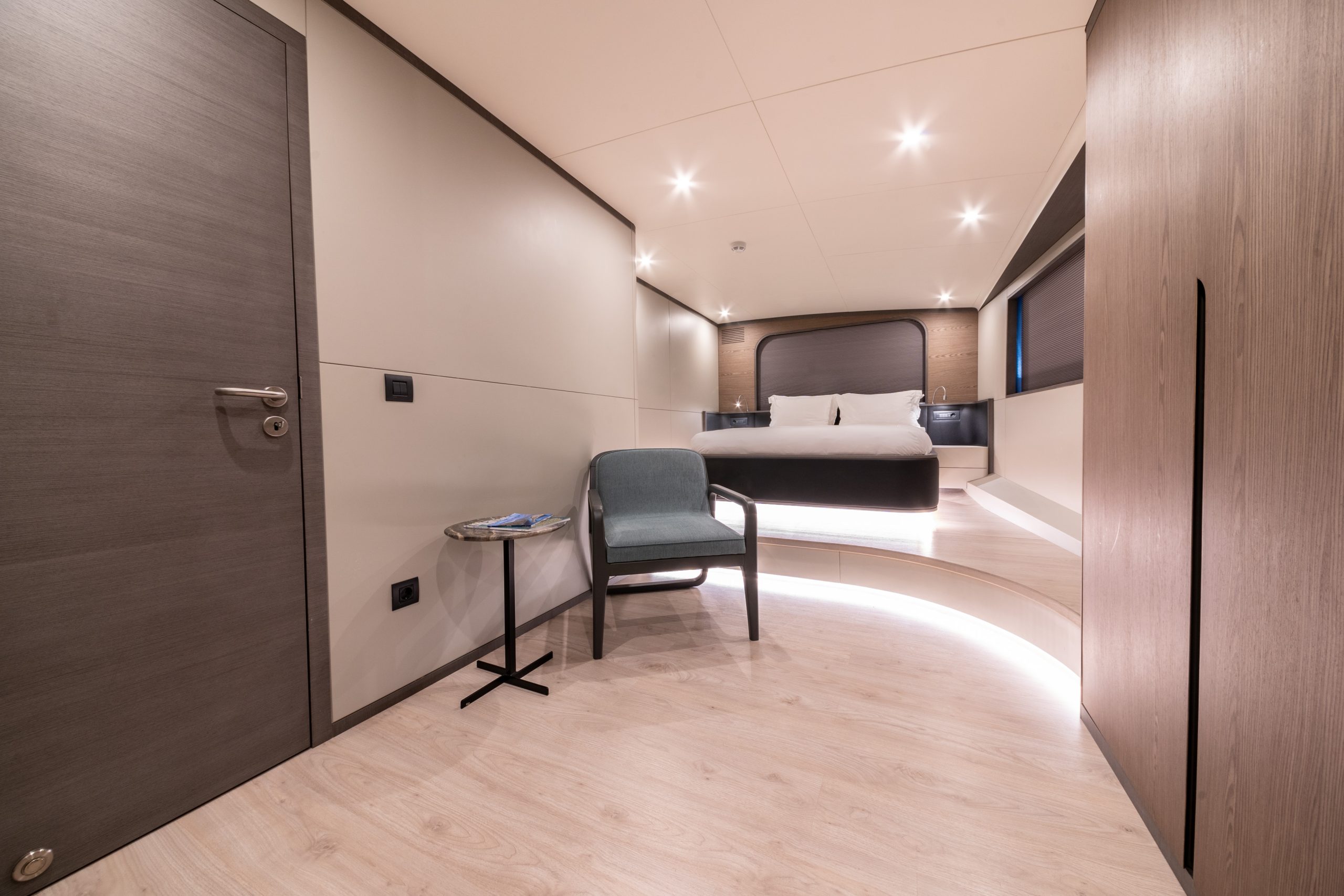 GOOD LIFE Modern, minimalist bedroom with wood flooring, a large bed with white bedding, a blue armchair beside a small round table, and soft recessed lighting creating a warm, inviting ambiance.