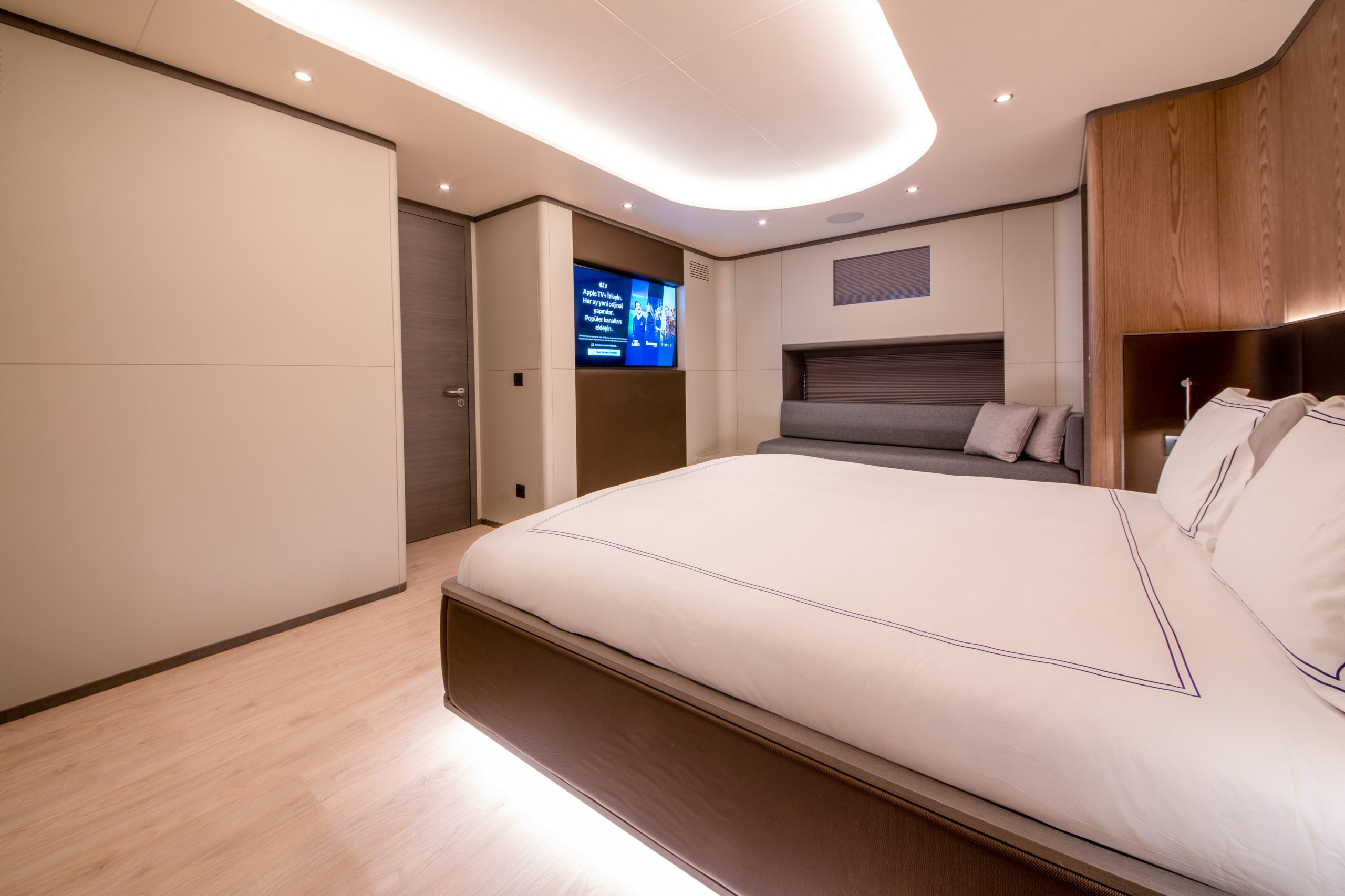 GOOD LIFE A modern, minimalist bedroom with a large bed, neutral colors, a sofa, wood accents, and a wall-mounted TV displaying a user interface. Recessed ceiling lights and under-bed lighting create a warm ambiance.
