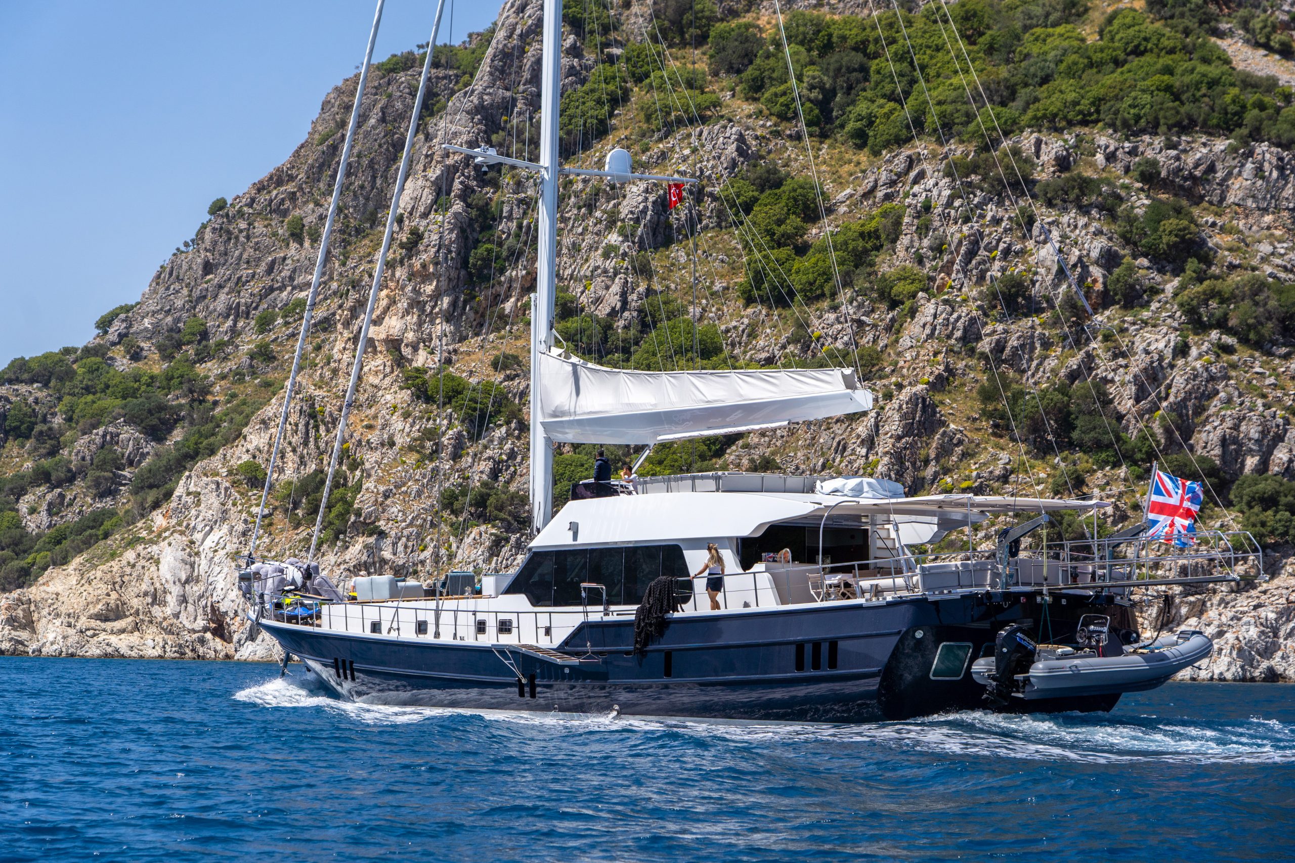 GOOD LIFE A sleek sailing yacht with a white canopy and British flag cruises on clear blue water near a rocky, green coastline under a bright sky.