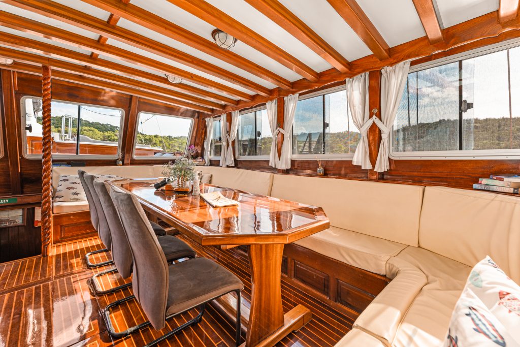 Interior of a wooden boat with large windows, beige cushioned seating along the wall, a polished wooden table, black chairs, and natural light highlighting the cozy, nautical ambiance.