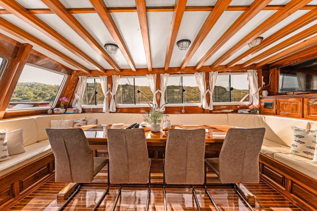 A bright, elegant yacht interior with a wooden dining table, six chairs, cream-colored cushioned seating, large windows with curtains, and decorative plants, offering a scenic outdoor view.