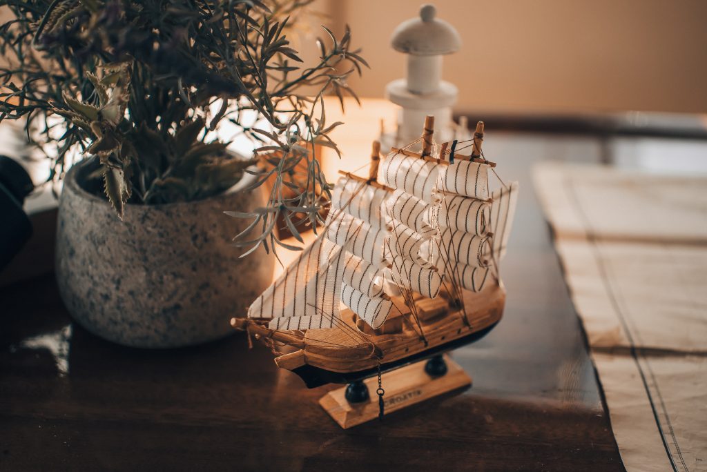 A detailed model ship with white sails sits on a polished wooden table beside a potted plant in a stone container and a white decorative object, with warm, soft lighting illuminating the scene.