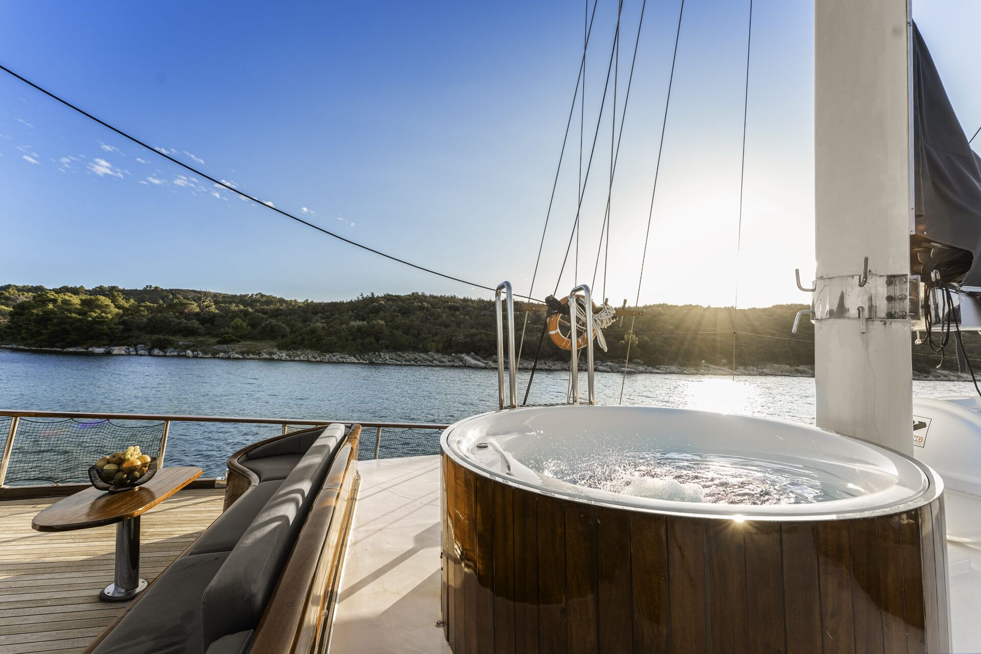 A hot tub on the deck of a yacht overlooks calm water and a scenic, tree-covered shoreline under a clear, sunny sky. Cushioned seating and a small table with fruit are nearby.