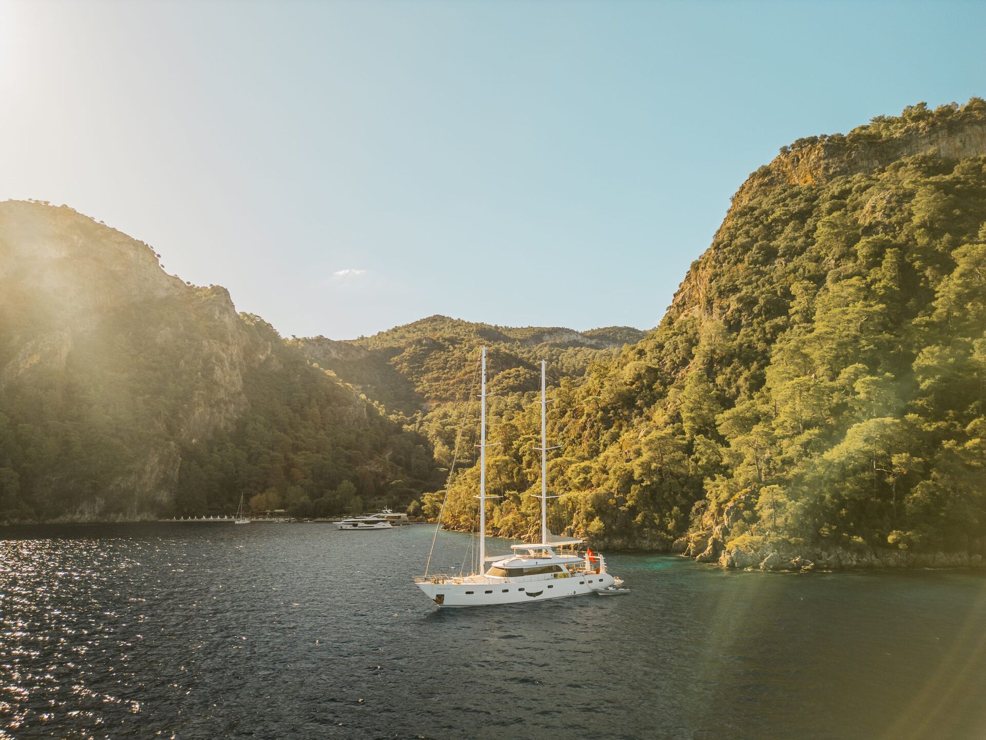 A white sailboat floats on clear blue water near a lush, green coastline with steep hills under a sunny, clear sky. Sunlight creates a warm glow, and other yachts for charter are visible in the background.