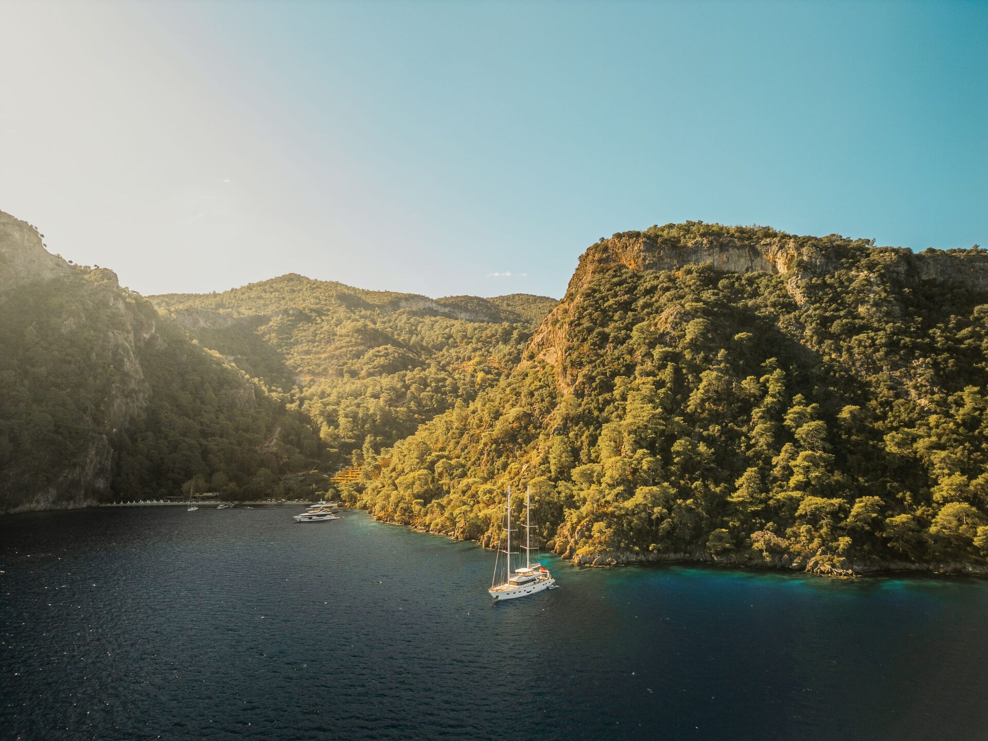 ANGELO III A sailboat floats on a calm, deep blue bay surrounded by green, forested hills under a clear sky, with sunlight illuminating part of the landscape.