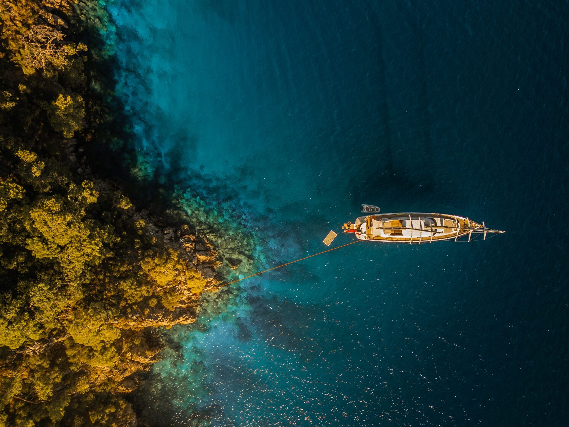 ANGELO III Aerial view of a boat anchored near a lush, green coastline with clear blue water. The boat is tied to the shore, and sunlight highlights both the water’s varying shades and the greenery along the shoreline.