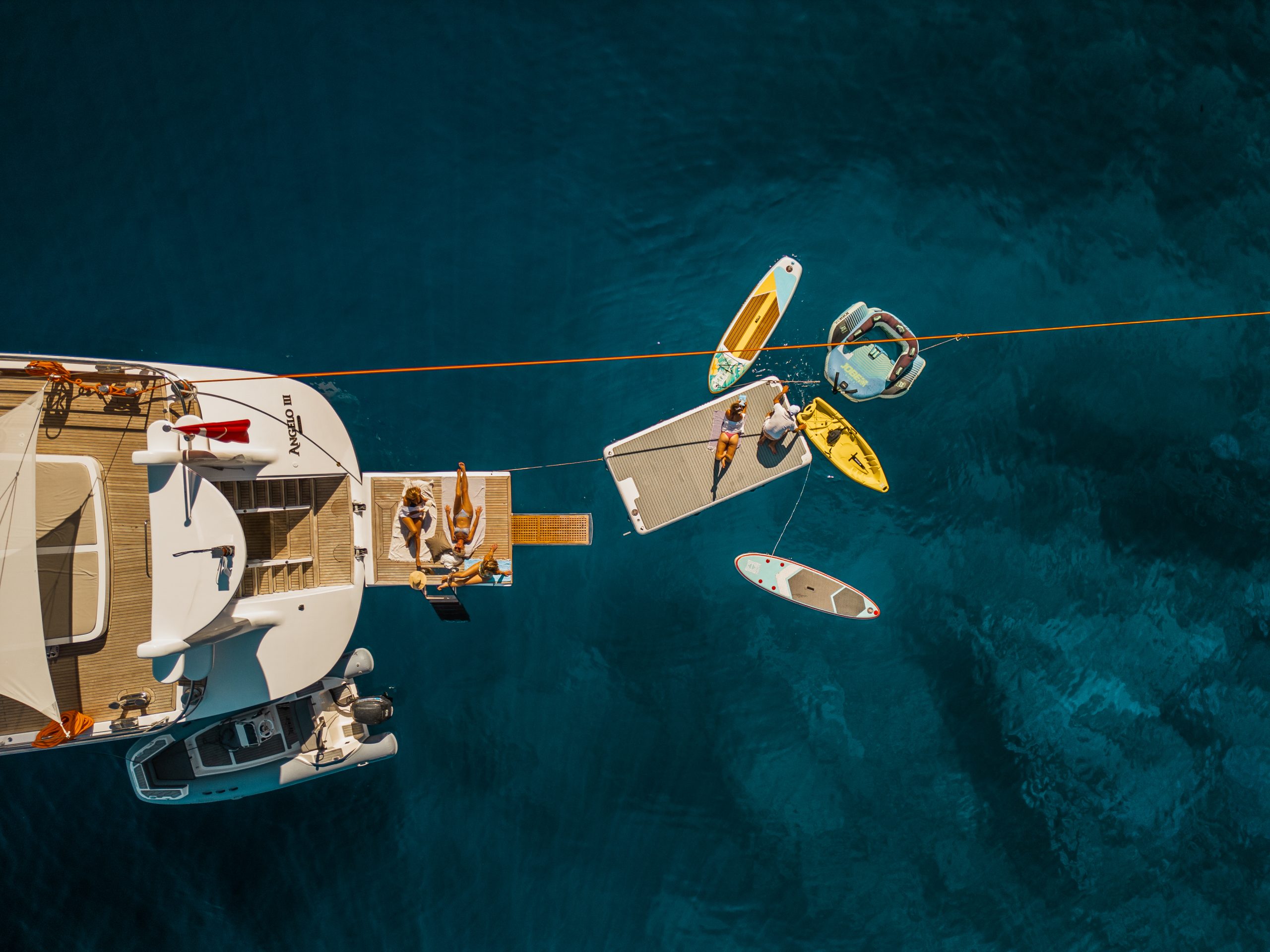 Aerial view of a yacht charter anchored in clear blue water, with people relaxing on floating platforms and various watercraft, including kayaks and paddleboards, surrounding the boat.