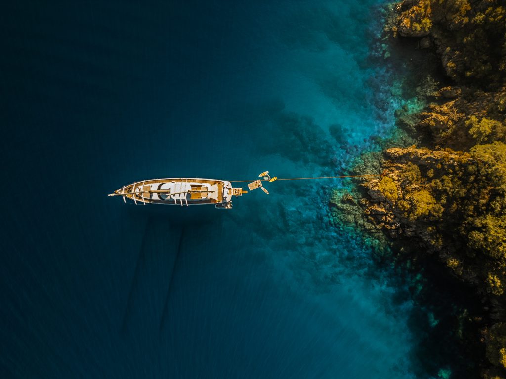 Aerial view of a yacht anchored in clear blue water near a rocky, yellow-brown coastline with visible underwater features—perfect for those seeking premium yachts for charter.