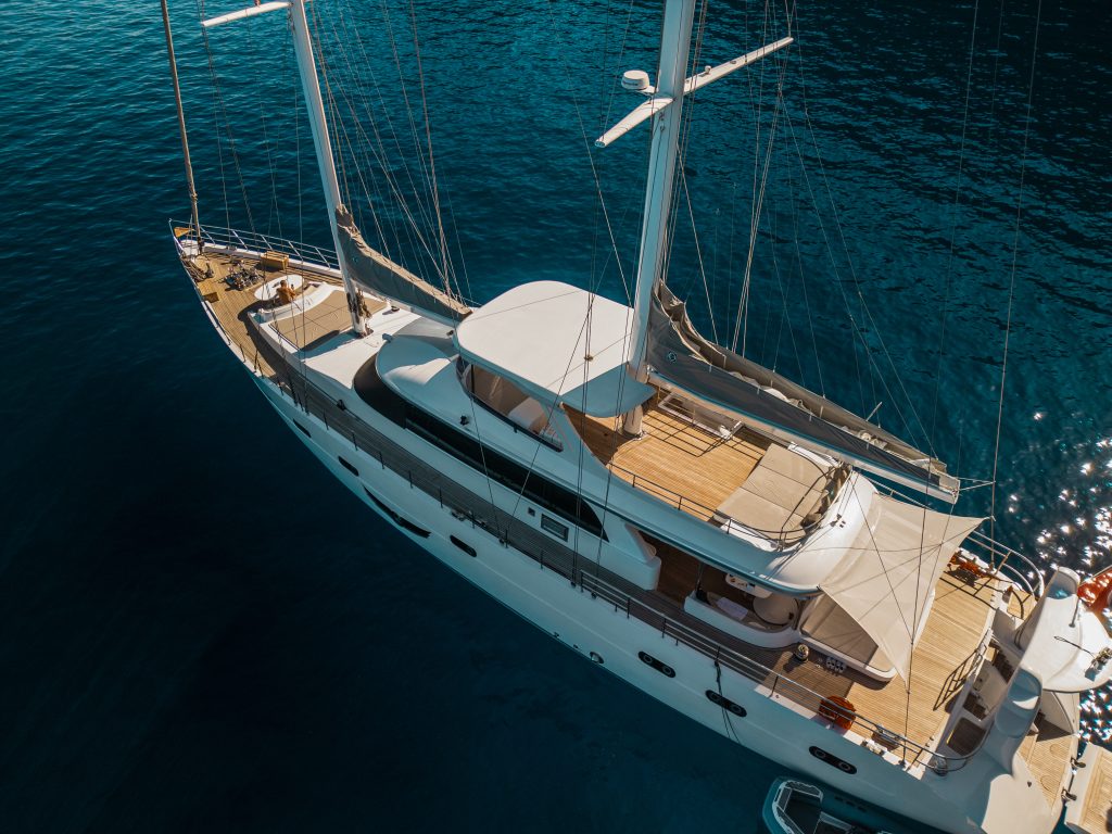 A large, luxurious white sailing yacht available for yacht charter is anchored in calm, deep blue water, viewed from above on a sunny day. The spacious deck features seating areas and sunbathing spaces.
