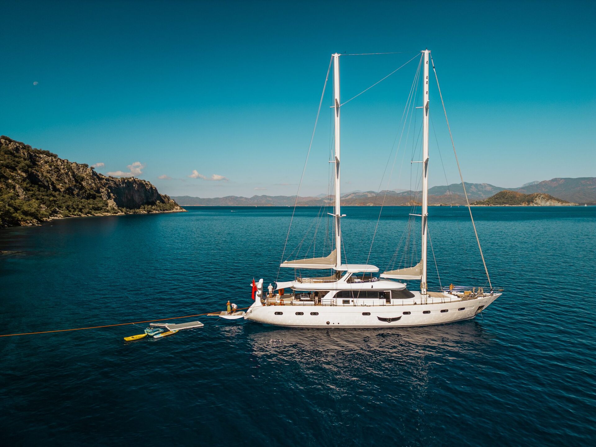 A large white yacht for charter is anchored in calm blue water near a rocky coastline, with people on deck and a small boat tied alongside under a clear sky.