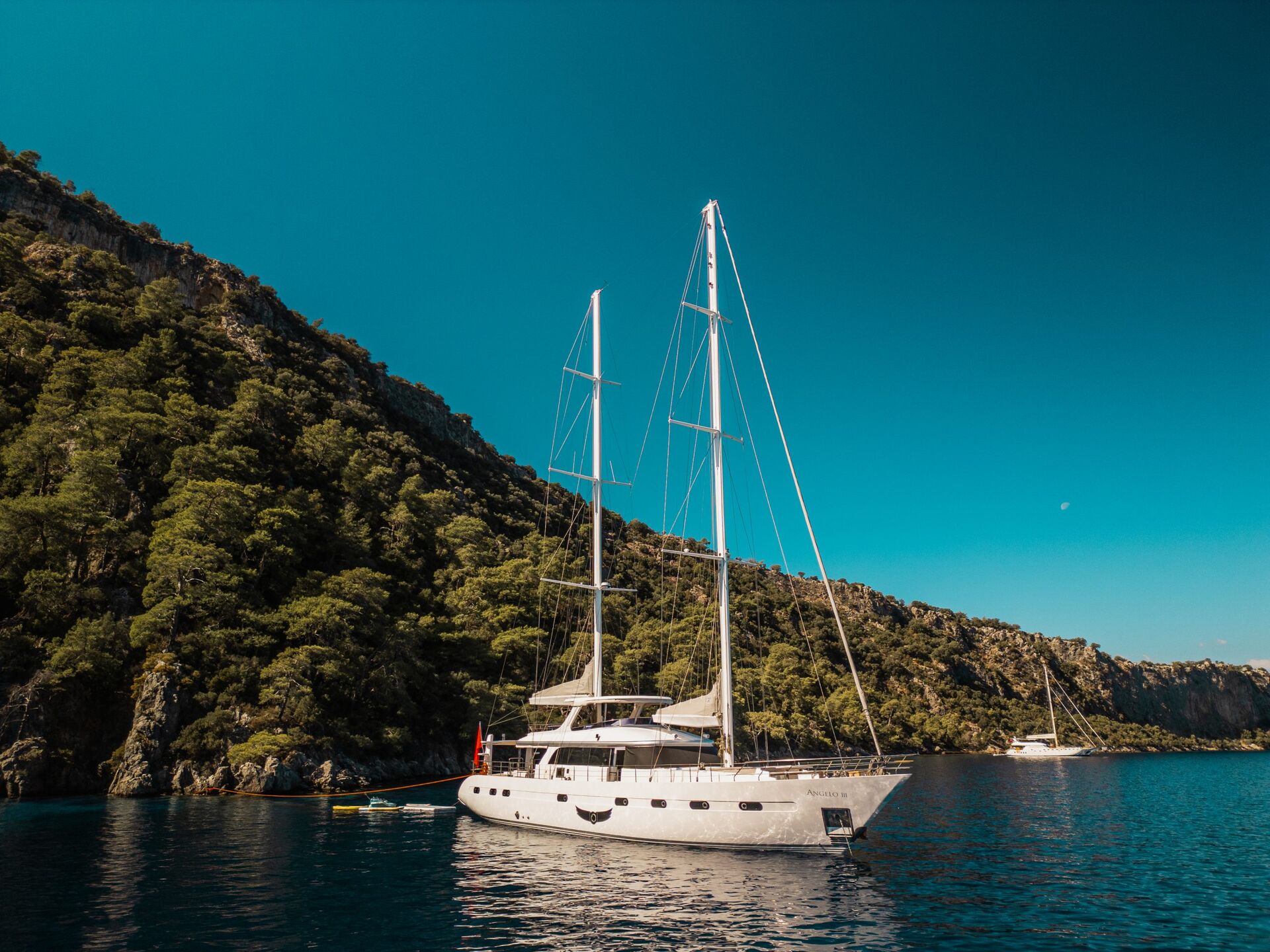 A large white sailboat, available for yacht charter, is anchored on calm blue water near a lush, tree-covered hillside under a clear blue sky. Another boat is visible in the distance.