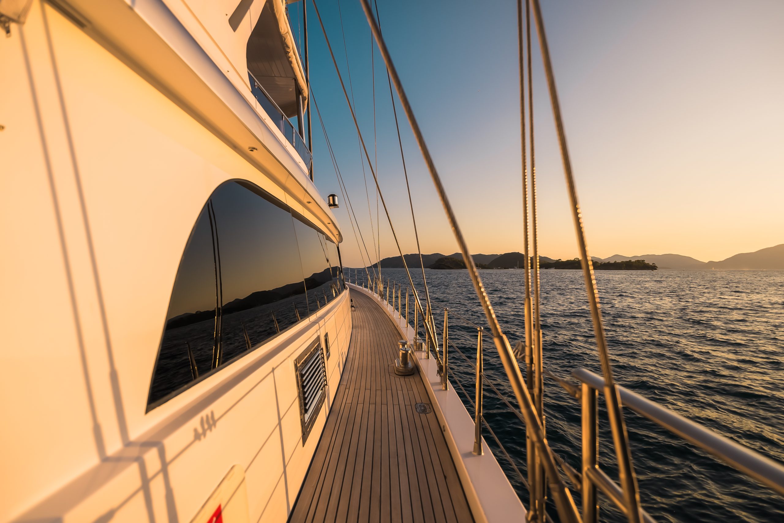 ANGELO III The deck of a yacht at sunset, with smooth wooden flooring and metal railings, sailing on calm water near distant mountains under a clear sky.