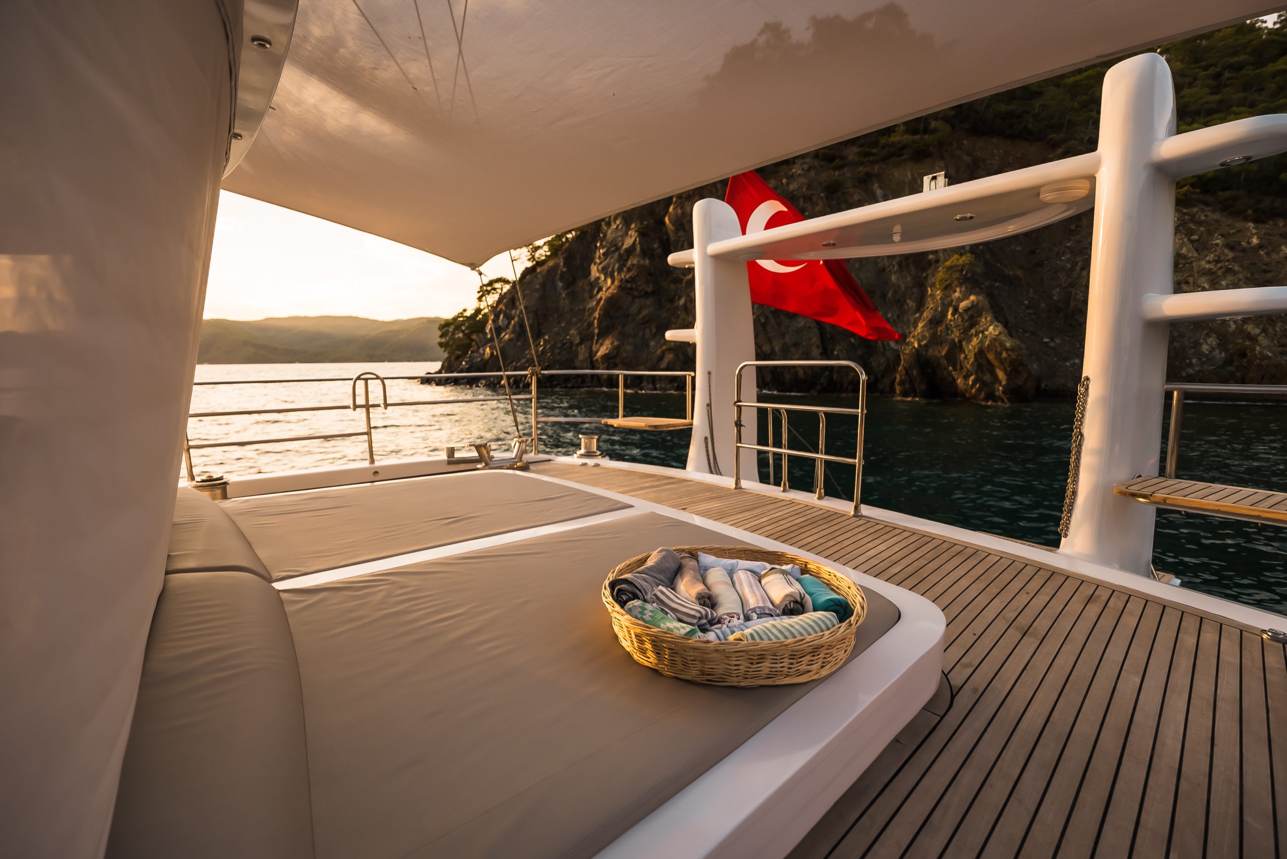 ANGELO III A cozy lounge bed with a basket of towels on the deck of a yacht, overlooking calm water and rocky cliffs at sunset, with a red Turkish flag in the background.