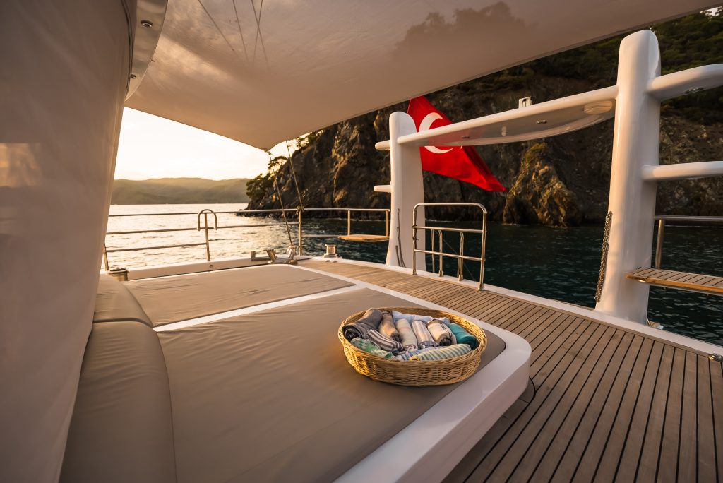 A sunlit yacht deck with a cushioned lounging area, a basket of towels, and a Turkish flag, overlooking calm water and rocky cliffs at sunset—an inviting scene perfect for your next yacht charter.