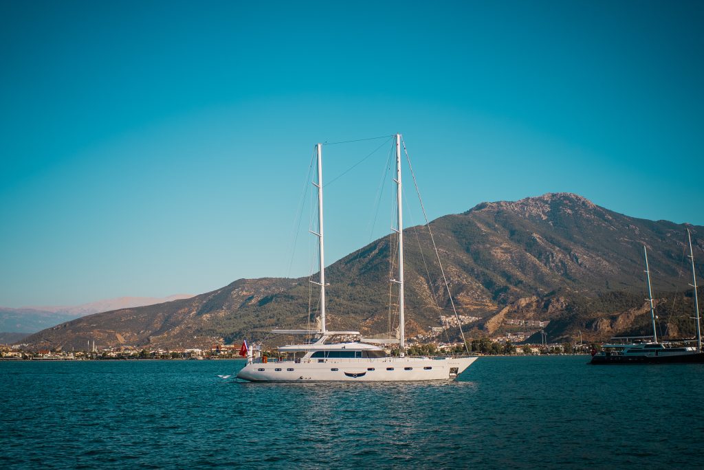 A white yacht with two masts floats on calm blue water, ideal for a yacht charter, with green, tree-covered mountains and a clear blue sky in the background. Another boat is partially visible to the right.