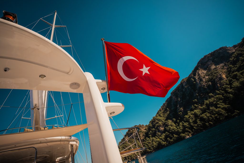 A Turkish flag waves on the back of a white yacht charter, with a rocky green hillside and deep blue water under a clear sky in the background.