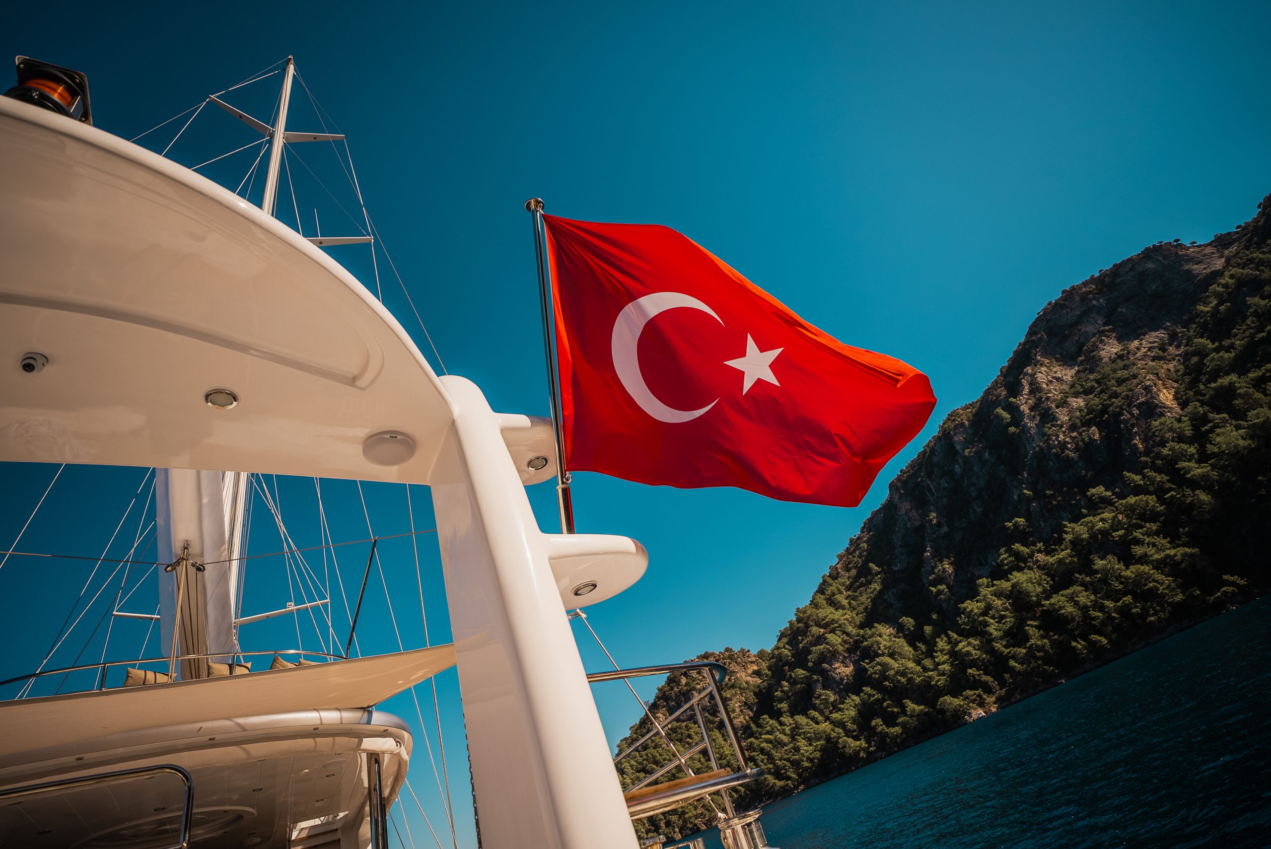A Turkish flag waves on the back of a white yacht charter, with a rocky green hillside and deep blue water under a clear sky in the background.