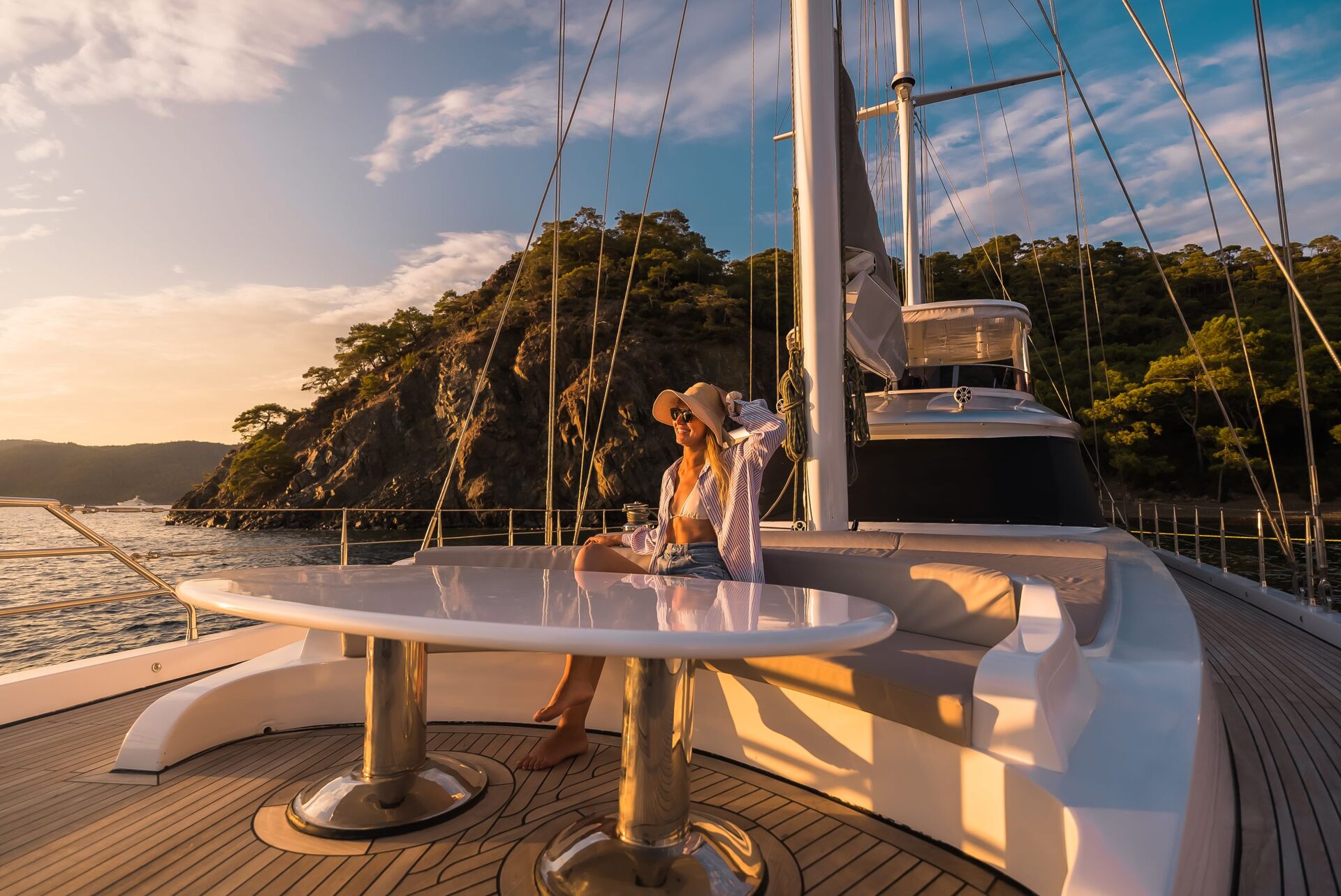 ANGELO III A woman wearing a hat and sunglasses relaxes on the deck of a yacht at sunset, with a drink in hand. The yacht is near a rocky, tree-covered shoreline under a partly cloudy sky.