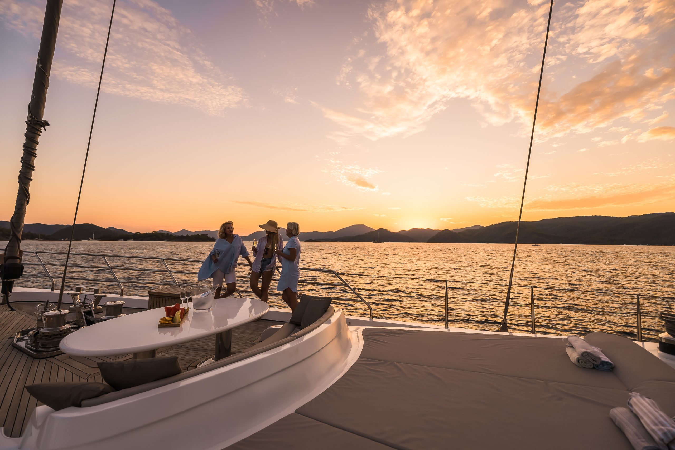 ANGELO III Three people in light clothing stand on the deck of a yacht at sunset, with drinks on a table nearby. The sun is setting over calm water and distant hills, casting a warm glow over the scene.