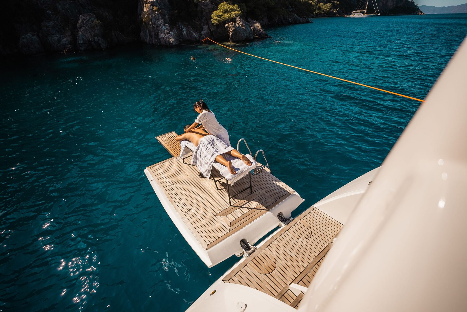 ANGELO III A person receives a massage on a platform at the back of a yacht, surrounded by turquoise water, with rocky cliffs and trees in the background.