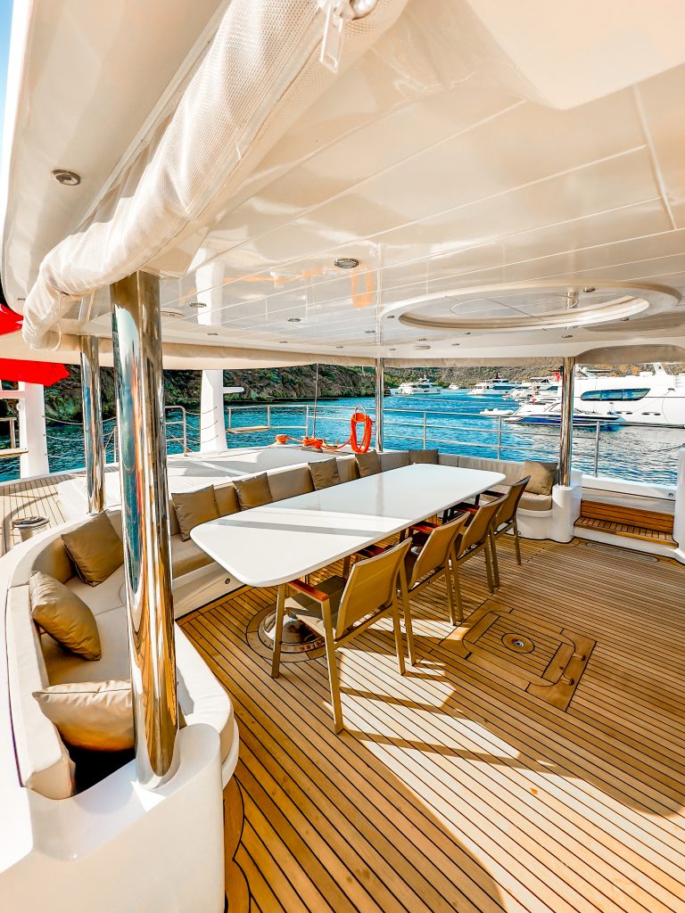 A luxurious yacht charter deck featuring a long white dining table with chairs, beige cushioned seating, polished wooden floor, and views of blue water with other boats and green hills in the background.
