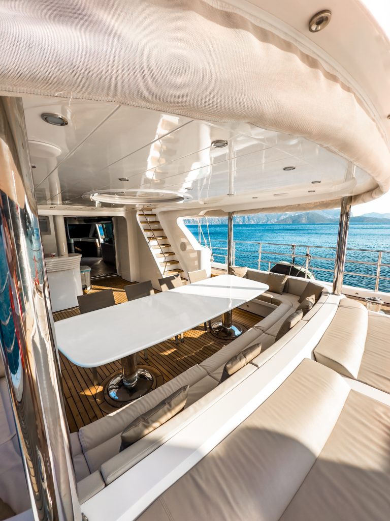 Outdoor dining area on a luxury yacht rental featuring a sleek white table, cushioned seating, and panoramic ocean views. Sunlight illuminates the modern deck, with stairs leading to an upper level and water visible in the background.
