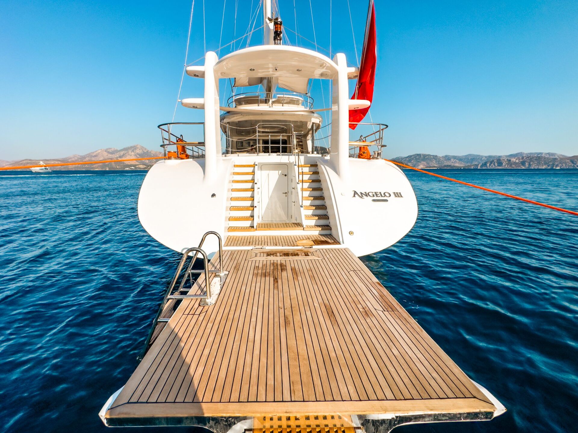 A luxurious white yacht named ANGELO III, available for yacht charter, is anchored in calm blue water. Viewed from the rear, its wooden swim platform extends toward the viewer under a clear sky and distant hills.