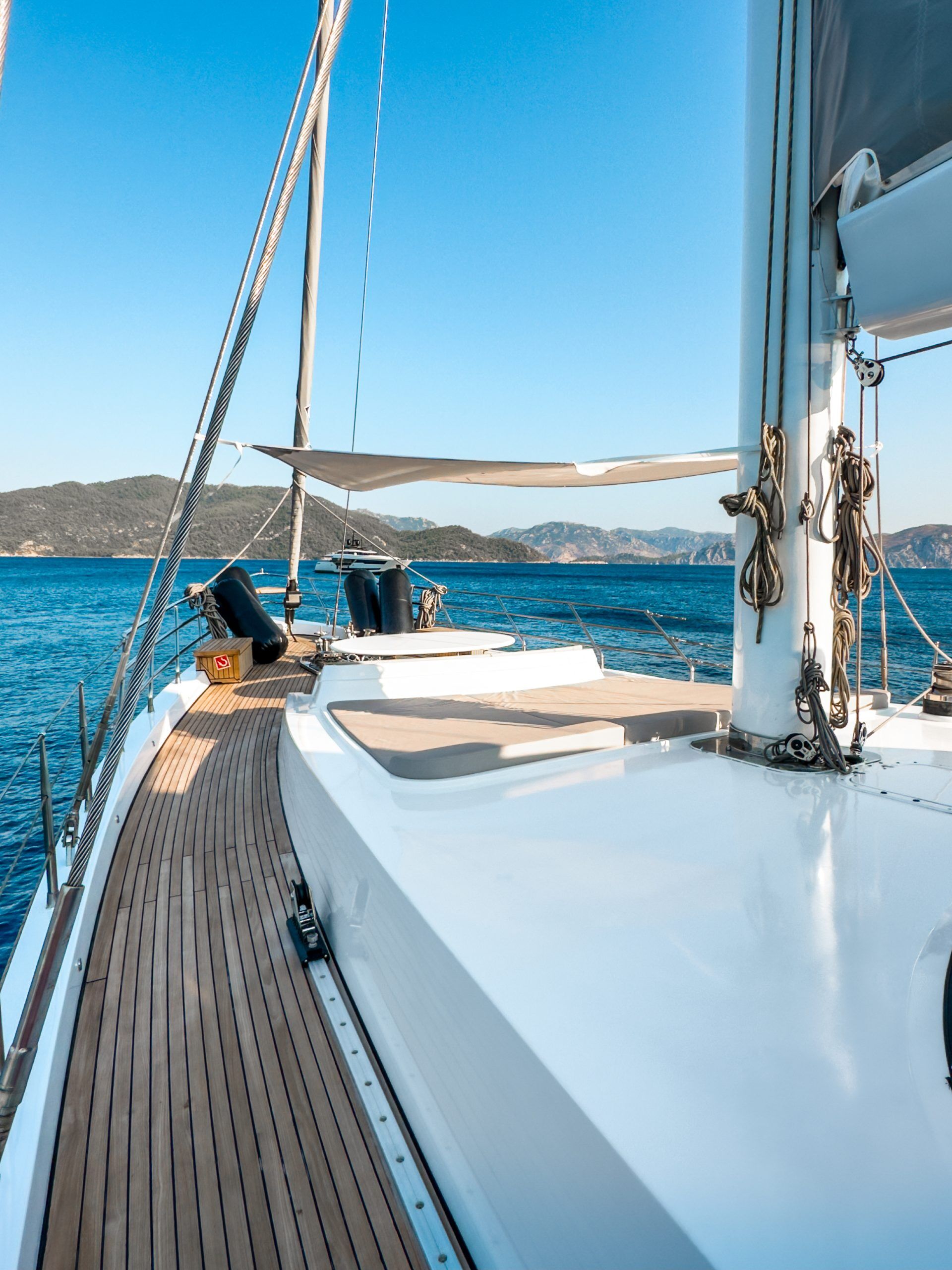 View from the deck of a sailboat with wooden flooring, calm blue sea, distant green mountains, and a clear sky—an idyllic scene perfect for those seeking a peaceful yacht charter experience.