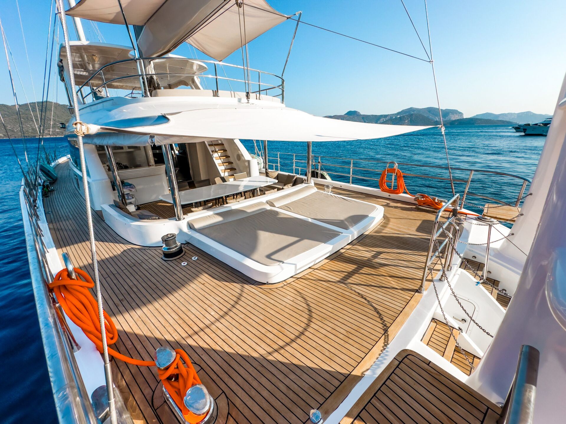 Luxury yacht deck with sun loungers, shaded areas, and wooden flooring, anchored on calm blue water near distant mountains under a clear sky. Perfect for a relaxing yacht rental, with orange lifebuoys and ropes visible on deck.